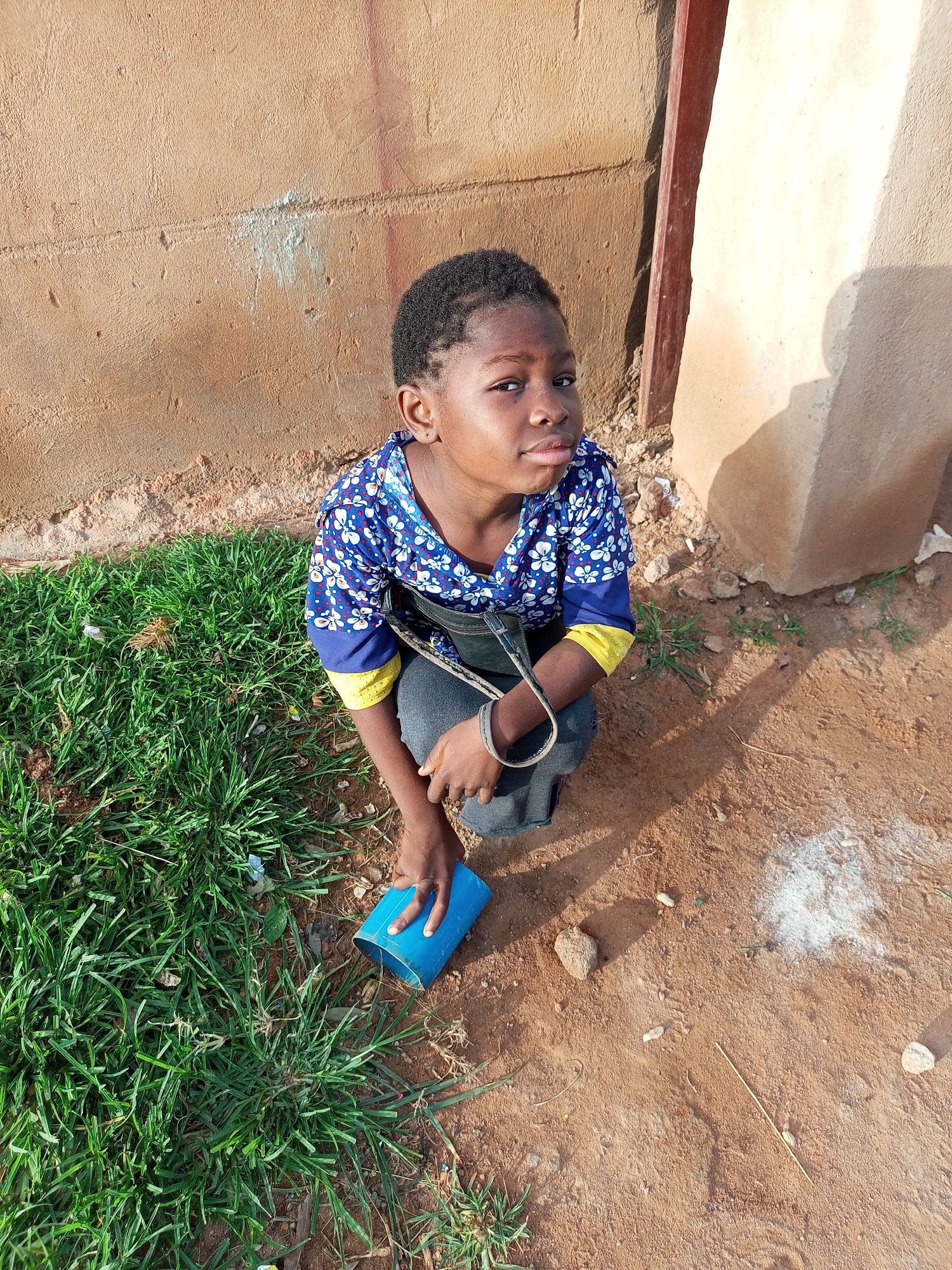 A young Black child kneels on the ground in front of a textured, tan-colored wall in Tempure, Nigeria. The child, with short, dark hair, wears a blue shirt with a white floral pattern and yellow accents on the sleeves. A dark strap, possibly from a bag, is draped across the child's chest. The child is looking directly at the camera with a neutral expression. To the left of the child is a patch of green grass. In front of the child, resting on the sandy ground, is a blue plastic cylinder. The ground is a mix of reddish-brown soil, small stones, and sparse dry grass. A vertical, dark reddish-brown wooden beam is visible on the right side of the image, attached to the wall. The lighting suggests it is daytime, with shadows cast on the wall.