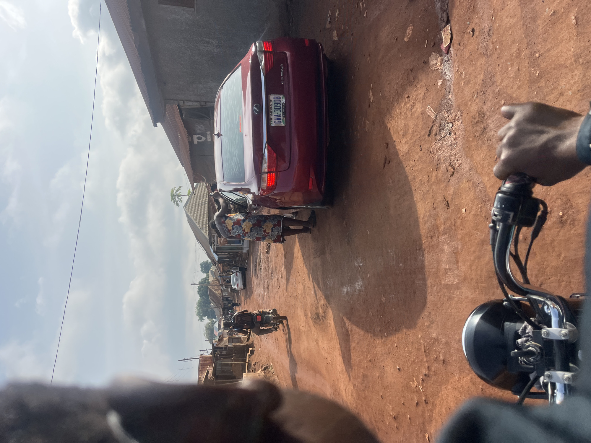 The image, taken from the vantage point of someone on a moving motorcycle, shows a street scene in what appears to be a densely populated area, likely on the outskirts of Abuja, Nigeria.
The road is unpaved, with a red-brown surface. On the left side of the road is a burgundy Lexus ES 350, partially blocking the road. Two individuals are standing near the car. Buildings line both sides of the road, consisting of corrugated metal structures and what appear to be informal shops or residences. A motorcycle is further down the road, and other vehicles and people are visible in the distance. The sky is partly cloudy. A license plate GWA-309DB.