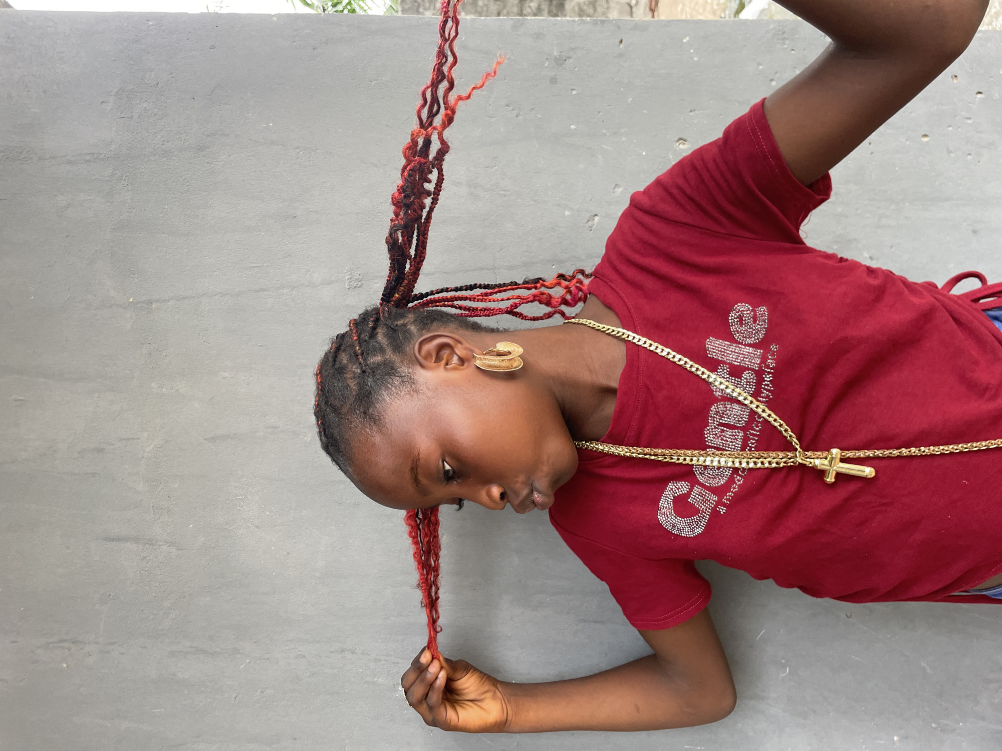 A close-up, slightly overhead shot captures a young person with dark skin, positioned at an angle against a plain light gray concrete wall. The person is leaning forward and downward, with their head turned to the left side of the frame, looking intently at their left hand. Their hair is styled in numerous small braids, predominantly black at the roots and transitioning to a vibrant red and black mix, with some longer, curly red strands extending upwards and others held in their hand.
They are wearing a short-sleeved, dark red t-shirt. The front of the t-shirt features the word "Guess" spelled out in sparkly, silver-colored rhinestones. Around their neck, a thick, gold-colored chain with a prominent gold crucifix pendant hangs. A large, ridged, gold-colored hoop earring is visible in their right ear. The left hand, which they are observing, gently holds a single curly red braid. The background is consistently a light gray concrete wall, with a hint of blurry green foliage and a lighter surface, possibly sky or another structure, at the top left, suggesting an outdoor setting in Badagry, Nigeria. The lighting indicates it is daytime.