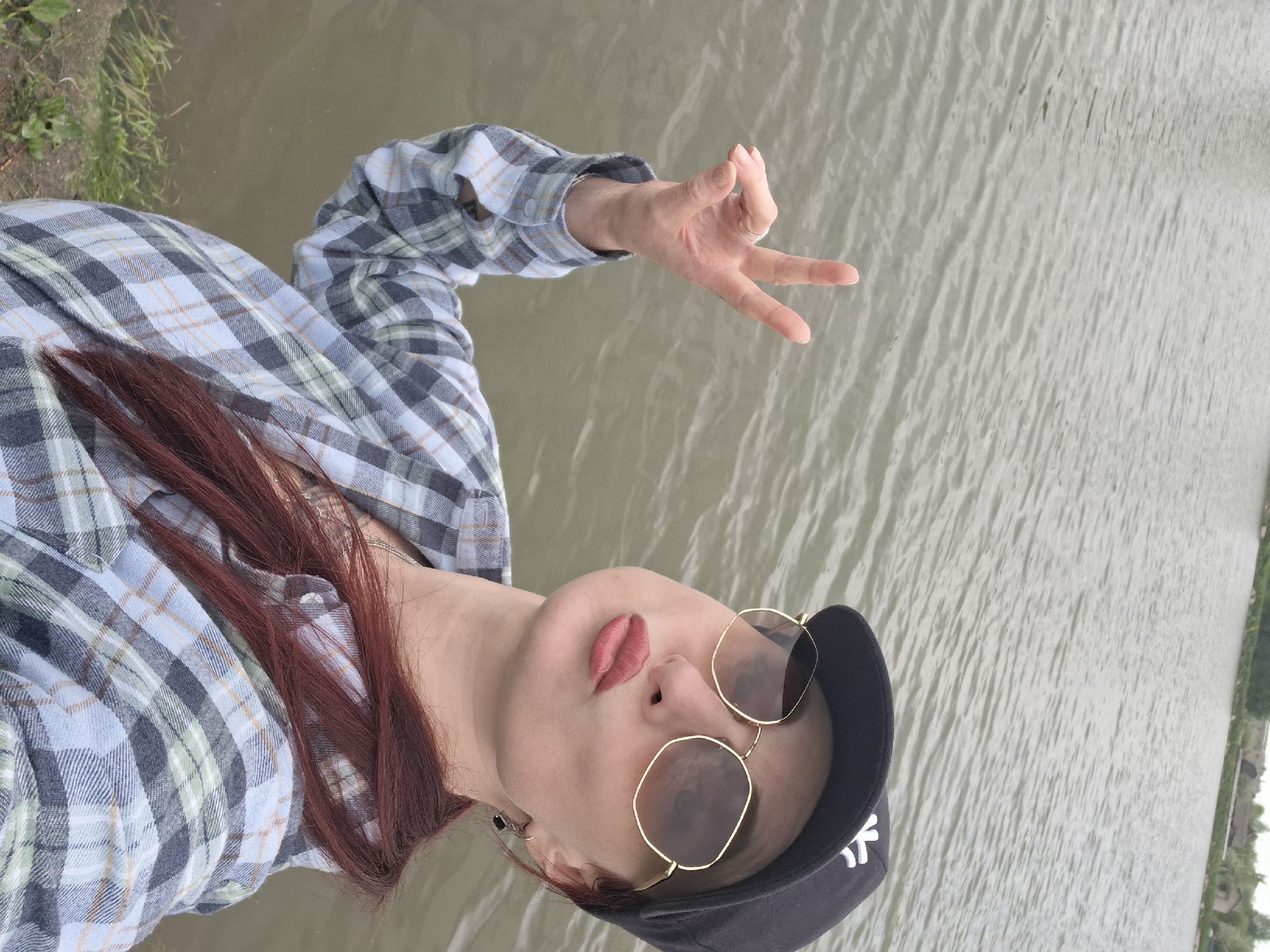 A woman is near water, possibly a lake, in Забалоцкі сельскі Савет, Belarus. She's wearing a blue and gray plaid shirt, a black baseball cap, and round gold-rimmed sunglasses. She has red hair and is making a hand gesture. The water is gray and rippled. Vegetation is visible on the distant shore.