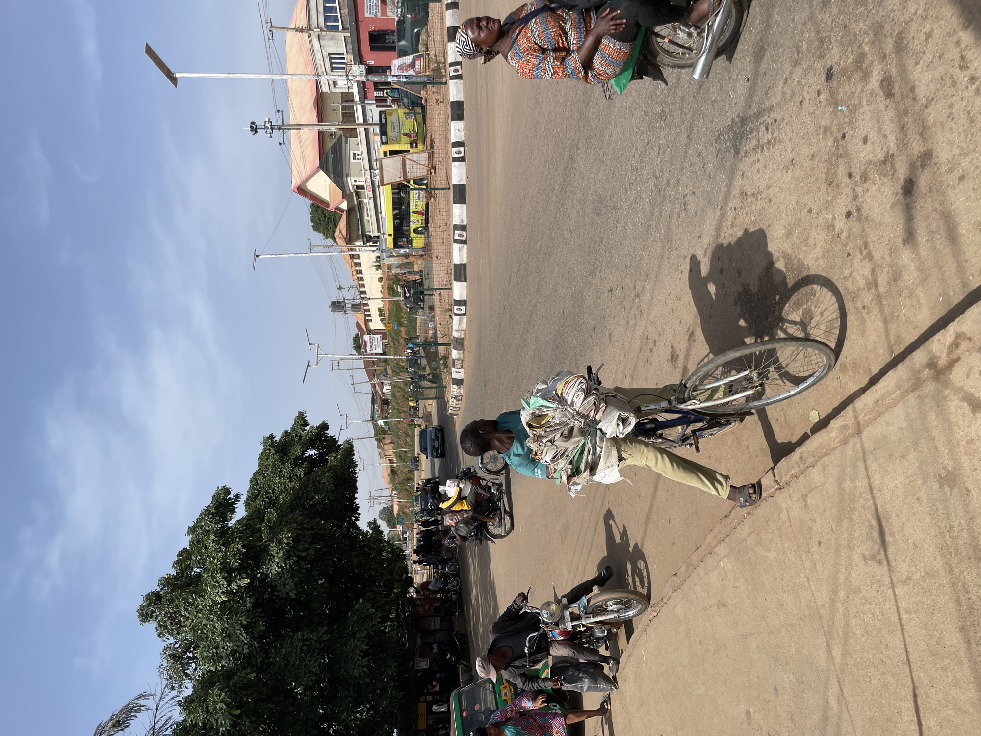The image depicts a street scene in Bukuru, Nigeria. In the foreground, a man is riding a bicycle laden with a large bundle of items, possibly textiles or papers. His shadow is visible on the road surface. Adjacent to the curb, a line of motorcycles is parked. Further down the street, there are various vehicles, including cars and buses. Buildings are visible along the street, and a line of utility poles extends into the distance under a blue sky. A woman in patterned clothing is seated on a motorcycle.