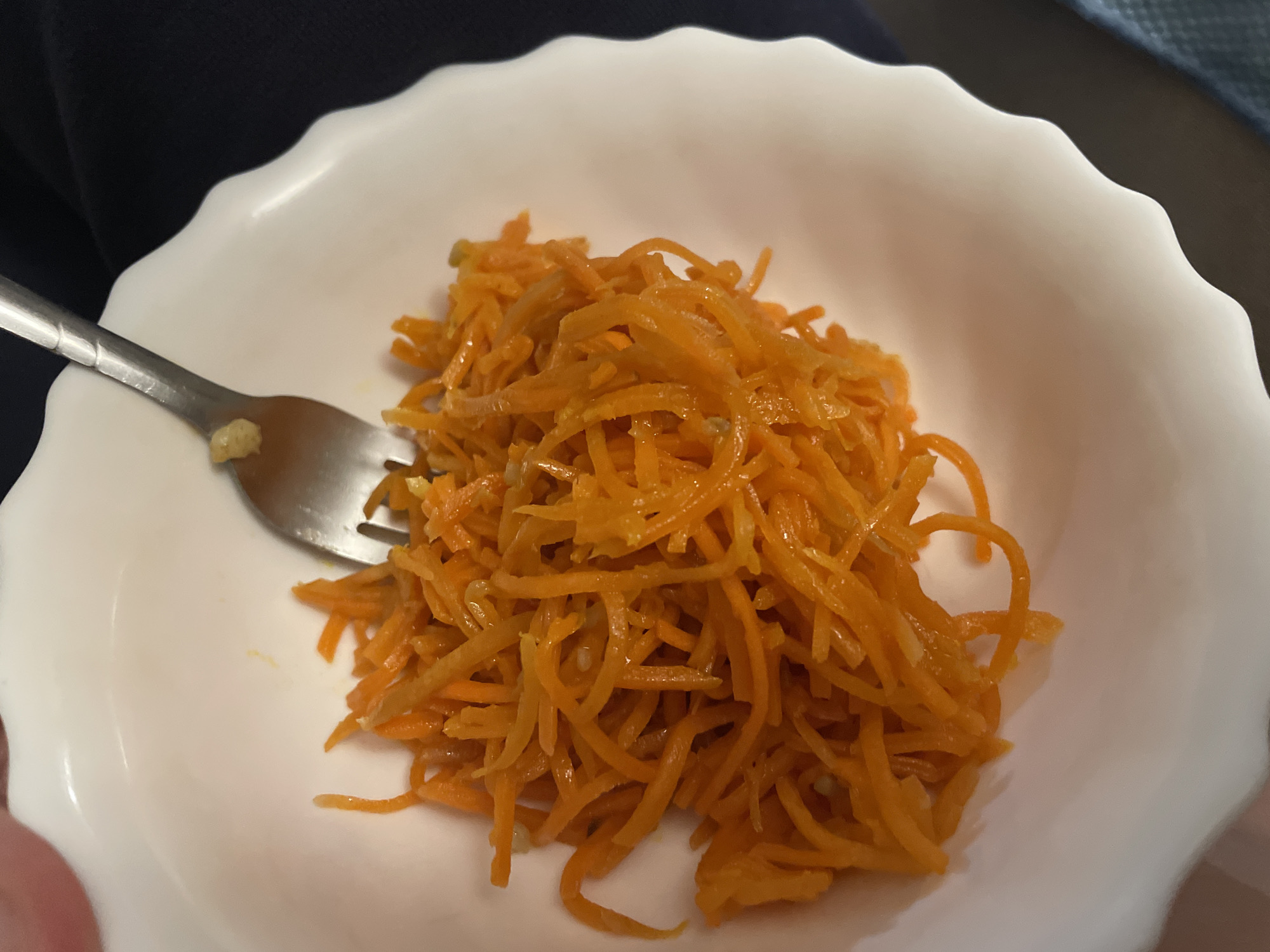 A close-up, overhead shot captures a white, fluted bowl filled with shredded carrots. A silver fork is positioned in the bowl, with a small portion of food on its tines. The background is dark and out of focus, suggesting an indoor setting. There is no visible information to determine the location in Minsk, Belarus.