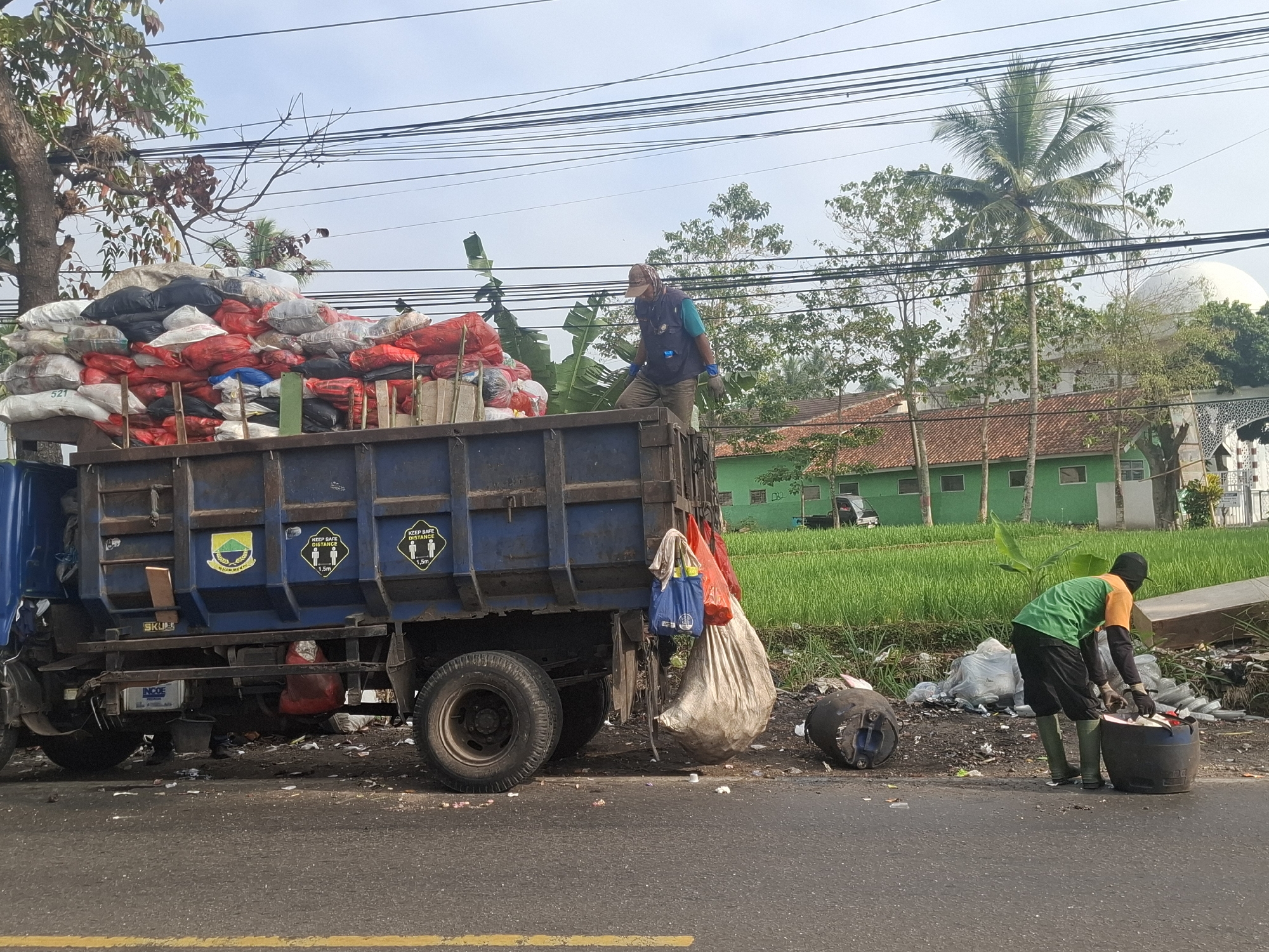 In Rancagoong, Indonesia, a blue truck is parked on the side of a road, its bed filled with numerous trash bags. The bags are of various colors, including red, white, and black. A man wearing a blue vest over a light blue shirt, a cap, and gloves is standing on the truck, seemingly engaged in sorting or arranging the trash. Next to the truck, on the side of the road, two men are working. One man, wearing a green shirt, black shorts, and green boots, is kneeling and appears to be emptying a large black barrel filled with trash. Another black barrel lies on its side nearby. The surrounding environment features a grassy field behind the truck and a building with a red-tiled roof. Overhead, a complex network of power lines is visible against a clear sky. Trees, including a palm tree, are scattered throughout the landscape. The road itself has a yellow line marking its edge.