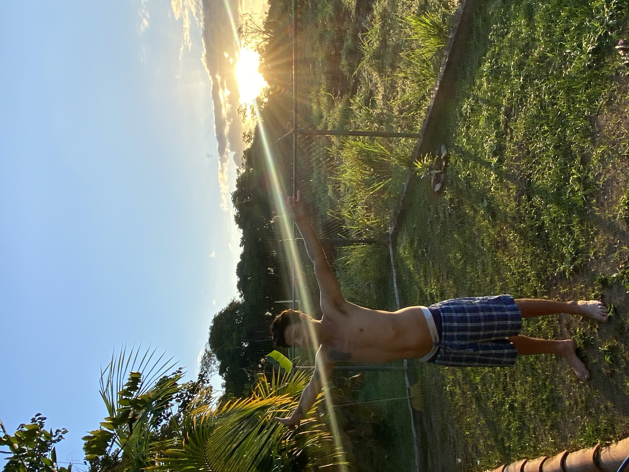 This image captures an outdoor scene in Los Velásquez, Venezuela, during late afternoon or early evening. A shirtless male individual, appearing to be a young adult, stands barefoot in the center-left of the frame, facing slightly right with both arms extended horizontally. He is wearing blue plaid shorts, with the white waistband of light-colored underwear visible above them. Tattoos are visible on his left bicep/shoulder area and his right forearm.
The ground is a grass-covered area with some visible patches of bare soil. In the foreground to the left, large green palm fronds are partially visible, and a yellow rake with a wooden handle rests on the ground behind the subject's left side. A metal chain-link fence spans horizontally across the midground, situated behind the individual and separating the immediate grassy area from a field of taller, overgrown green vegetation. A small, light-green plant surrounded by a few rocks is present in the grass near the fence, to the right of the subject.
The background features a dense line of trees along the distant horizon. The setting sun is prominent on the right side of the image, low in the sky behind scattered clouds, emitting strong golden light and distinct lens flares that cross the frame. The sky above is clear blue with wispy clouds near the sun.