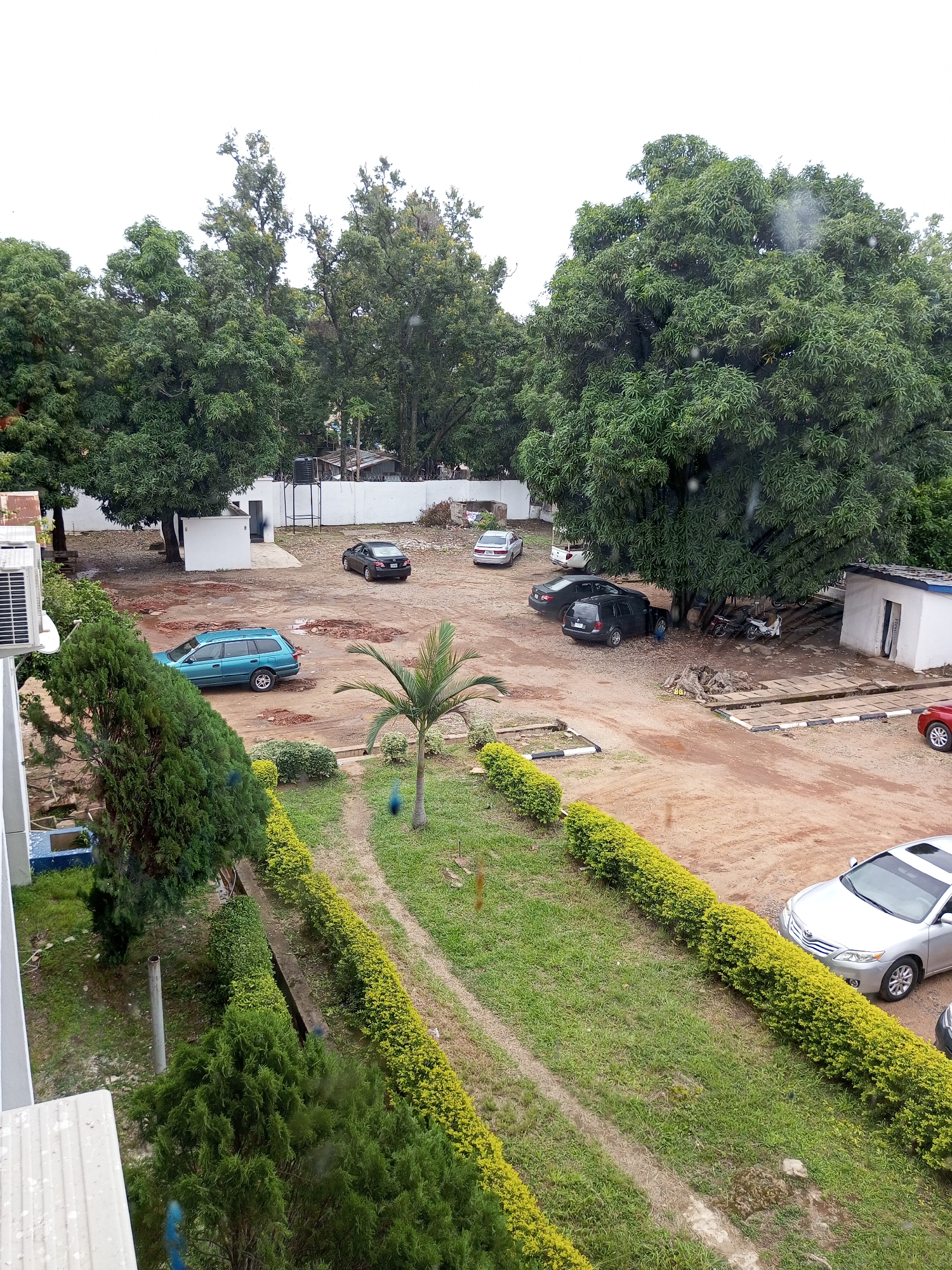 This is an aerial shot of a parking area in Jos, Nigeria. The scene depicts a mix of nature and human-made structures, with large, leafy trees dominating the background and a well-maintained lawn with hedges in the foreground. The parking lot itself appears to be dirt, with some puddles indicating recent rain.
Several vehicles are parked in the lot, including a teal station wagon, a black sedan, a dark SUV, a silver sedan, and a red car. There are also a few motorcycles parked under the shade of a large tree. A small, white building with a blue trim is visible on the right side of the frame, and a portion of another building with an air conditioning unit is visible on the far left.
The time of day is unclear, but the overcast sky suggests it might be late morning or afternoon, possibly during a cloudy or rainy period. The overall impression is one of a functional outdoor space within a tropical or sub-tropical environment, likely a commercial or residential complex given the presence of multiple vehicles and buildings. No people are clearly visible in the image. No text is discernible.