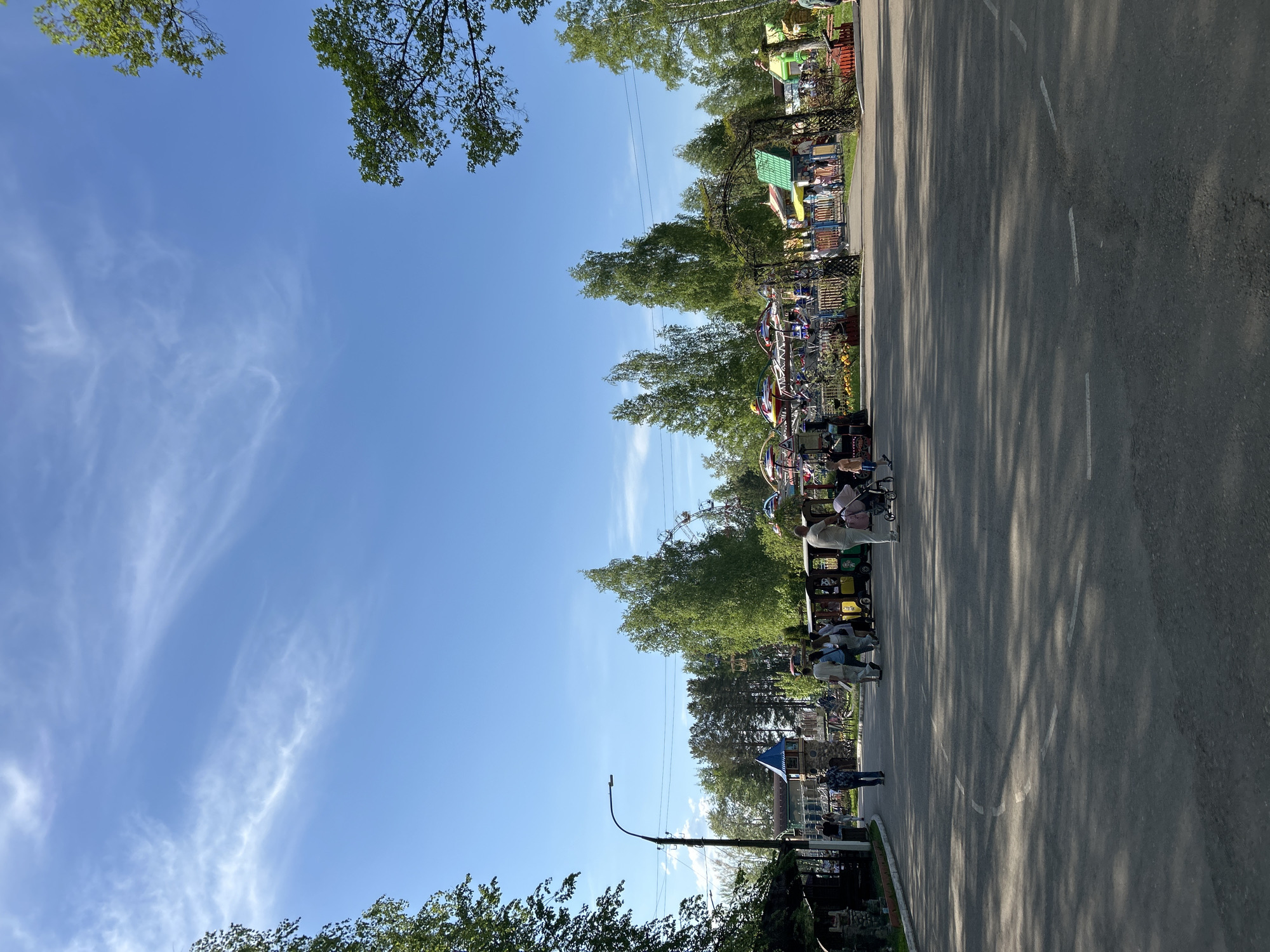 This is an outdoor shot taken on a bright sunny day in the city of Chaikovsky, Russia, likely during warm weather. It shows a roadside view within what appears to be an amusement park or recreational area. A wide asphalt road runs diagonally through the image, casting shadows from the trees lining the roadside. On the left side of the road, there are amusement park rides, some of which are visible behind the trees. There are several people visible; some seem to be walking along the road, and others appear to be riding on a small amusement park train. The sky above is clear with a few wispy clouds. The trees are lush with green leaves. There's a light pole and some structures (possibly buildings) on the far left, providing context for the scene being an urban leisure area.