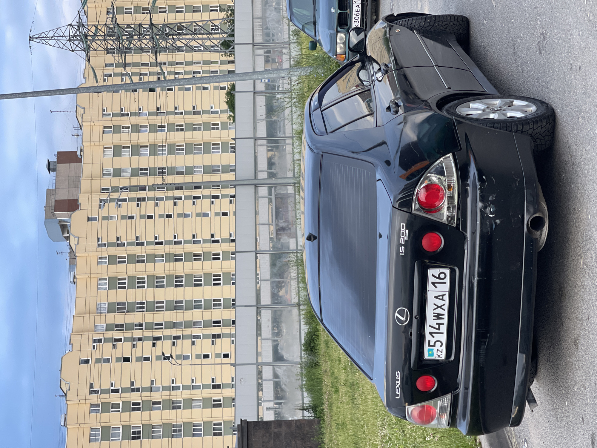 A black Lexus IS 200 is parked on an asphalt road. The car has a license plate that reads "KZ 514WXA 16". In the background, there is a tall, multi-story apartment building with a yellow facade and green trim. A large metal electrical pylon stands to the right of the building. There is also a green BMW parked to the right of the Lexus. The sky is cloudy.