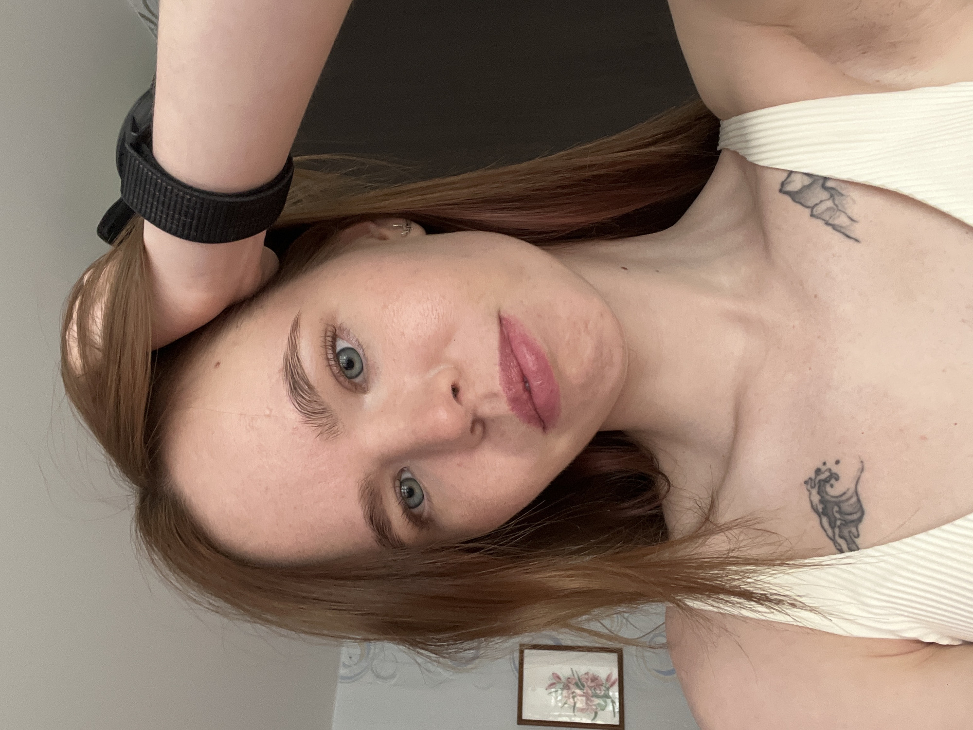 A woman with red hair, blue eyes, and fair skin is posing for a selfie. She is wearing a white, ribbed tank top and a black fitness tracker on her wrist. She has two visible tattoos, one on her neck and one on her chest. The background is a wall with a framed floral picture. The photograph was likely taken indoors. No evidence suggests this photograph was taken in Chaikovsky, Russia.