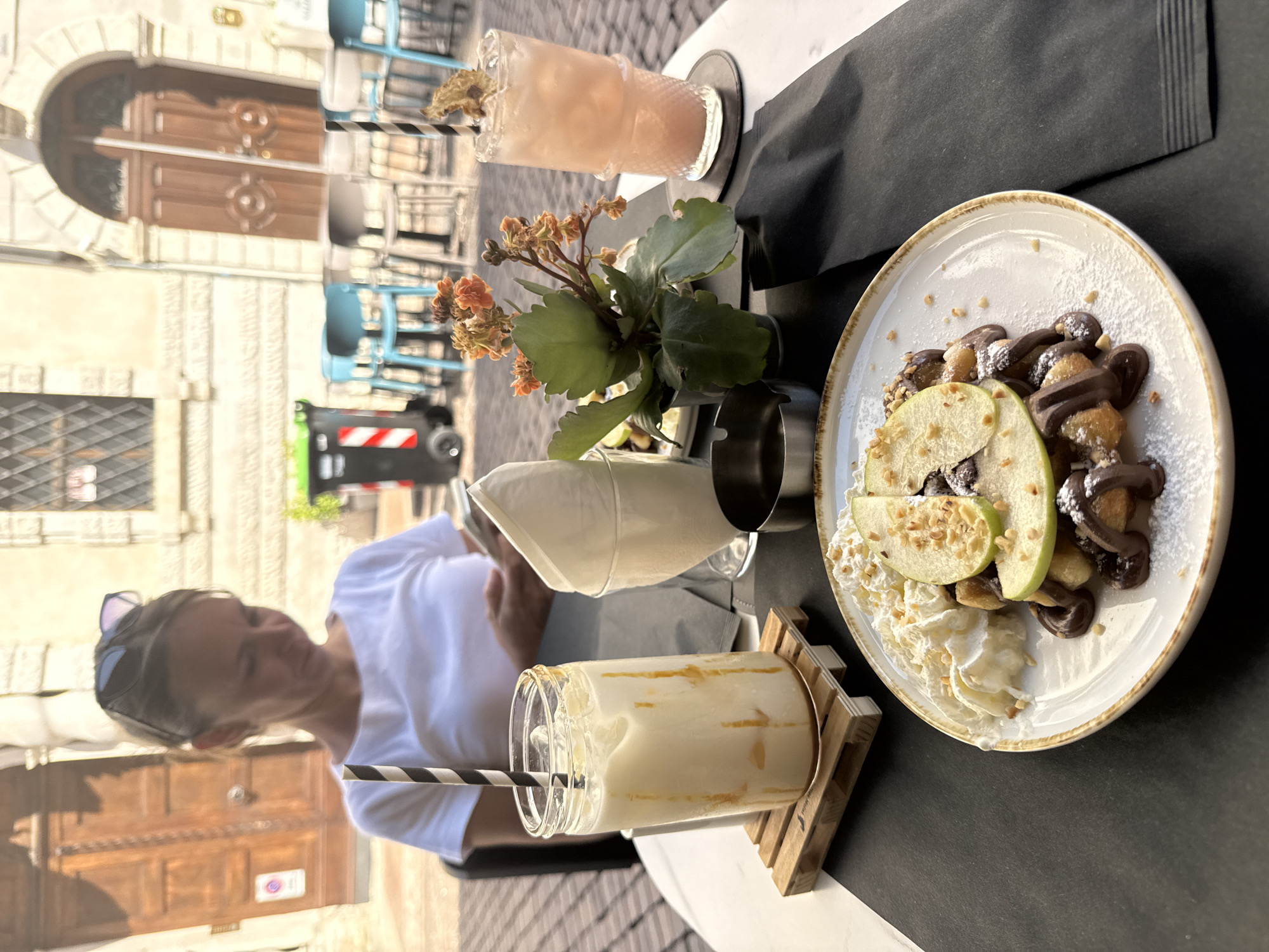 A woman is seated at an outdoor cafe table in Trento, Italy, looking down at her phone. In front of her is a plate of waffles with chocolate and whipped cream, a glass of a white drink with caramel drizzle, and a pink cocktail. A small potted plant with orange flowers sits between the drinks. The background shows a building with ornate wooden doors and windows, and outdoor seating with blue chairs. A trash bin is visible in the distance.