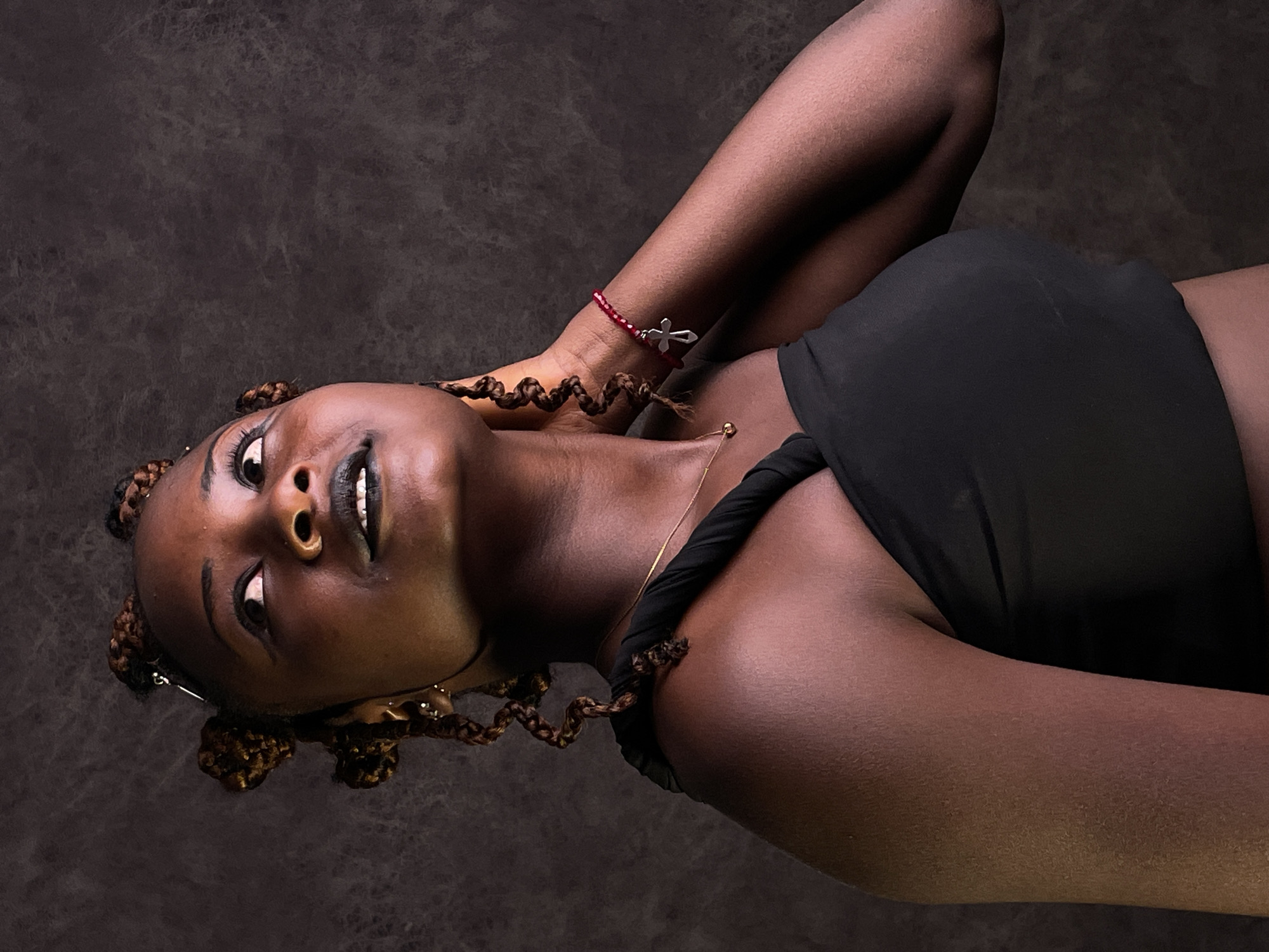 A medium shot depicts a woman lying on a dark, textured surface. She is dark-skinned with brown braided hair styled in buns. She wears black lipstick and a black top. A red beaded bracelet with a silver cross charm is visible on her arm. A thin gold necklace is around her neck. The location of the image cannot be determined from the visual information provided.