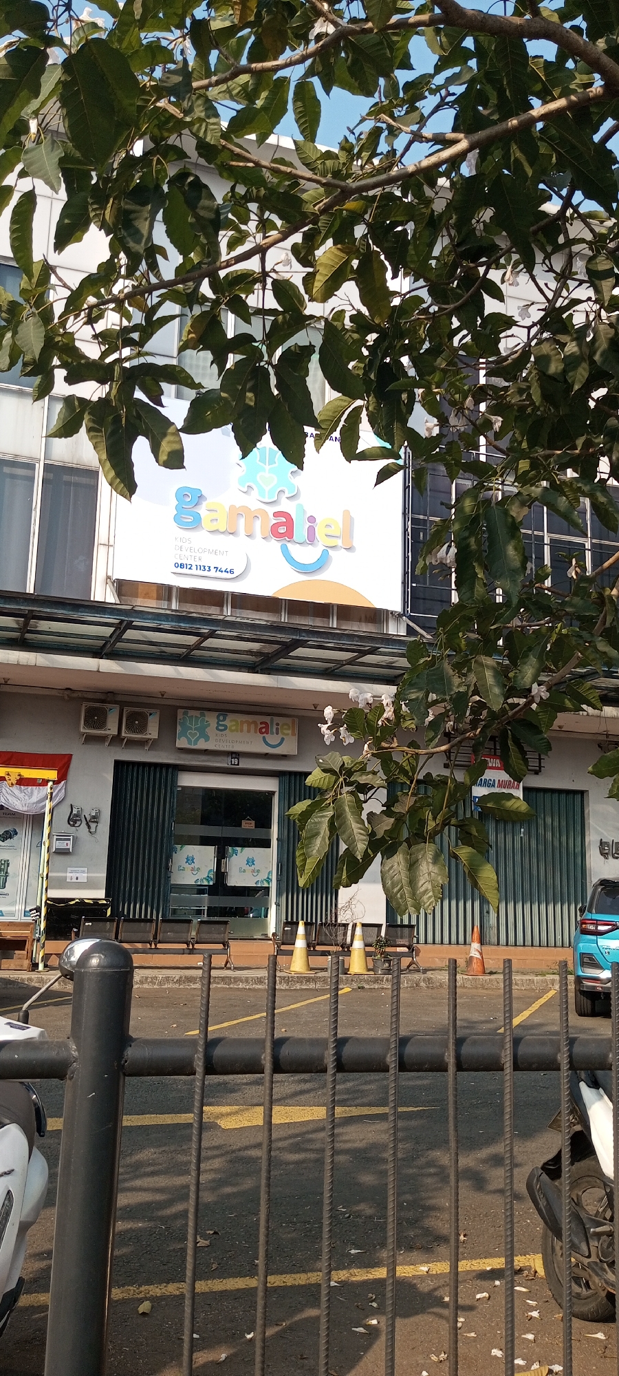 A medium-shot view captures the facade of a building adorned with large green leaves and branches from a nearby tree. The building features a white sign with the text "gamaliel KIDS DEVELOPMENT CENTER" in colorful lettering, accompanied by a phone number, "0812 1133 7446." Below the sign, green metal roll-up doors are partially open, revealing the entrance to the center. Two air conditioning units are mounted on the wall above the doors. In front of the building, several chairs are arranged on a tiled area, and traffic cones are placed along the pavement. A black metal fence with vertical bars runs across the foreground, partially obscuring a parking lot with yellow lines. A white motorcycle is visible on the left side of the frame, and a blue car is parked on the right. The scene is bathed in sunlight, casting shadows on the ground.