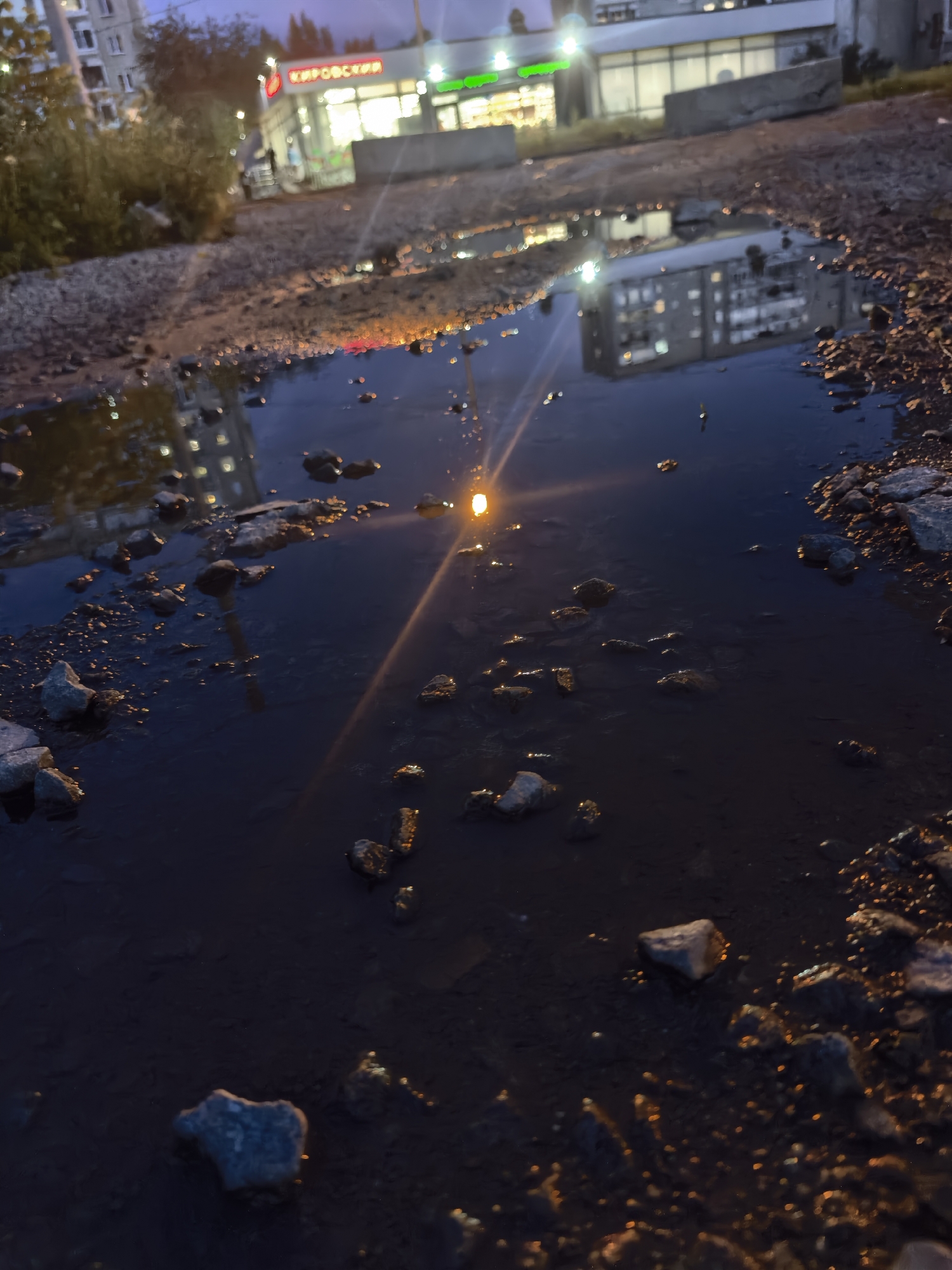 This is a low-angle, outdoor photograph taken at dusk. The foreground is dominated by a large, murky puddle reflecting the surrounding environment. The water is dark, with many small rocks and debris visible beneath its surface and along its edges. The reflection in the puddle shows buildings and lights, suggesting an urban or semi-urban setting.
In the background, a building with illuminated windows and signage is visible. The signage appears to be in Cyrillic script. To the left of this building, there are trees or bushes. The sky is dark, indicating evening or nighttime. The overall lighting is dim, with artificial lights from the building and a single, bright light source casting a lens flare or glare on the water's surface. This light source appears to be a streetlamp or a similar fixture, positioned to the right and slightly behind the viewer, casting a long reflection on the puddle.
The scene appears to be a somewhat neglected or underdeveloped area, with rough ground and a significant puddle, possibly after rain. No people are clearly visible in the image, though there might be indistinct figures in the background near the illuminated building. The mood is quiet and somewhat somber, characteristic of a twilight or evening scene in an urban fringe. The reflection in the puddle is a prominent feature, mirroring the built environment in the dark water. The text on the building appears to read "КИРОВСКИЙ".