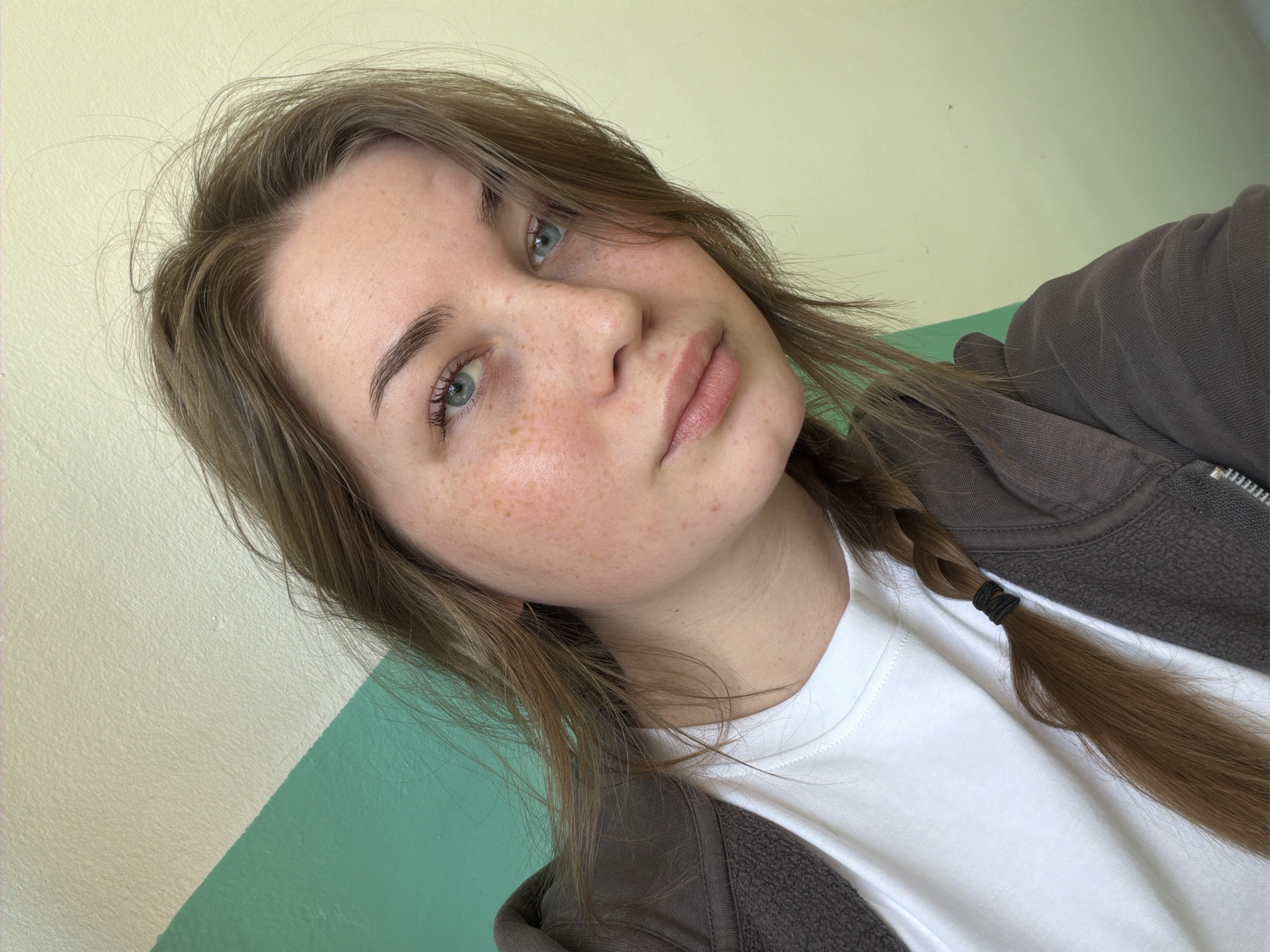 A young woman is posing for a selfie. She has fair skin with freckles, light brown hair partially styled in a braid, and blue eyes. She is wearing a white t-shirt and a brown zip-up jacket. The background is a wall painted in two colors, a light yellow or cream color and a green color. There is no information to determine the location as the city of Chaikovsky, Russia.