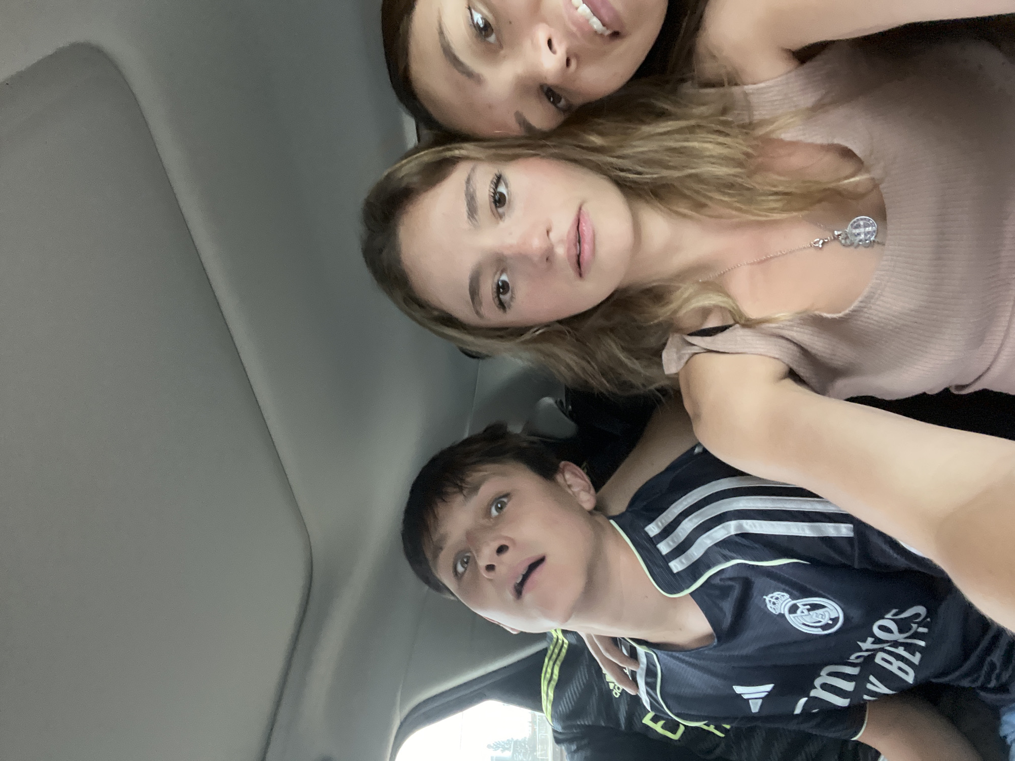 Three people are visible in the image, taken from a low angle, likely inside a vehicle. The vehicle's interior is gray.
The person closest to the camera is a young male with dark hair, wearing a black and white Adidas jersey. The jersey displays the "Real Madrid" logo.
Above him is a young woman with light brown hair, wearing a light pink top and a silver necklace.
The third person, partially visible at the top, is a young woman with dark hair.
The exact location within San Bernardino Tlaxcalancingo, Mexico, cannot be determined from the image.