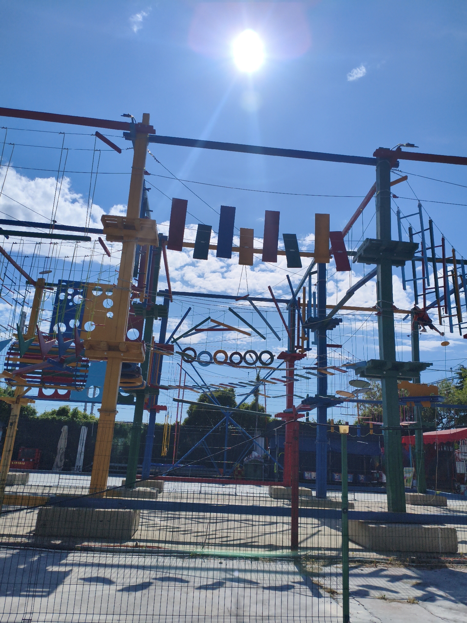 The image shows an outdoor ropes course under a bright, sunny sky with some clouds. The ropes course consists of various platforms, suspended bridges, nets, and obstacles made of wood and rope, colored in bright hues of yellow, red, blue, and green. The structures are tall and elaborate, forming a complex network. In the background, there are trees and some buildings, including what appears to be a red canopy structure. A person is visible on one of the suspended elements of the course, wearing a red harness. The ground beneath the course is paved, and a wire fence surrounds the area. The sun is high in the sky and appears to be creating lens flare in the image. This scene is located in Sochi, Russia, at an adventure park or similar recreational facility.