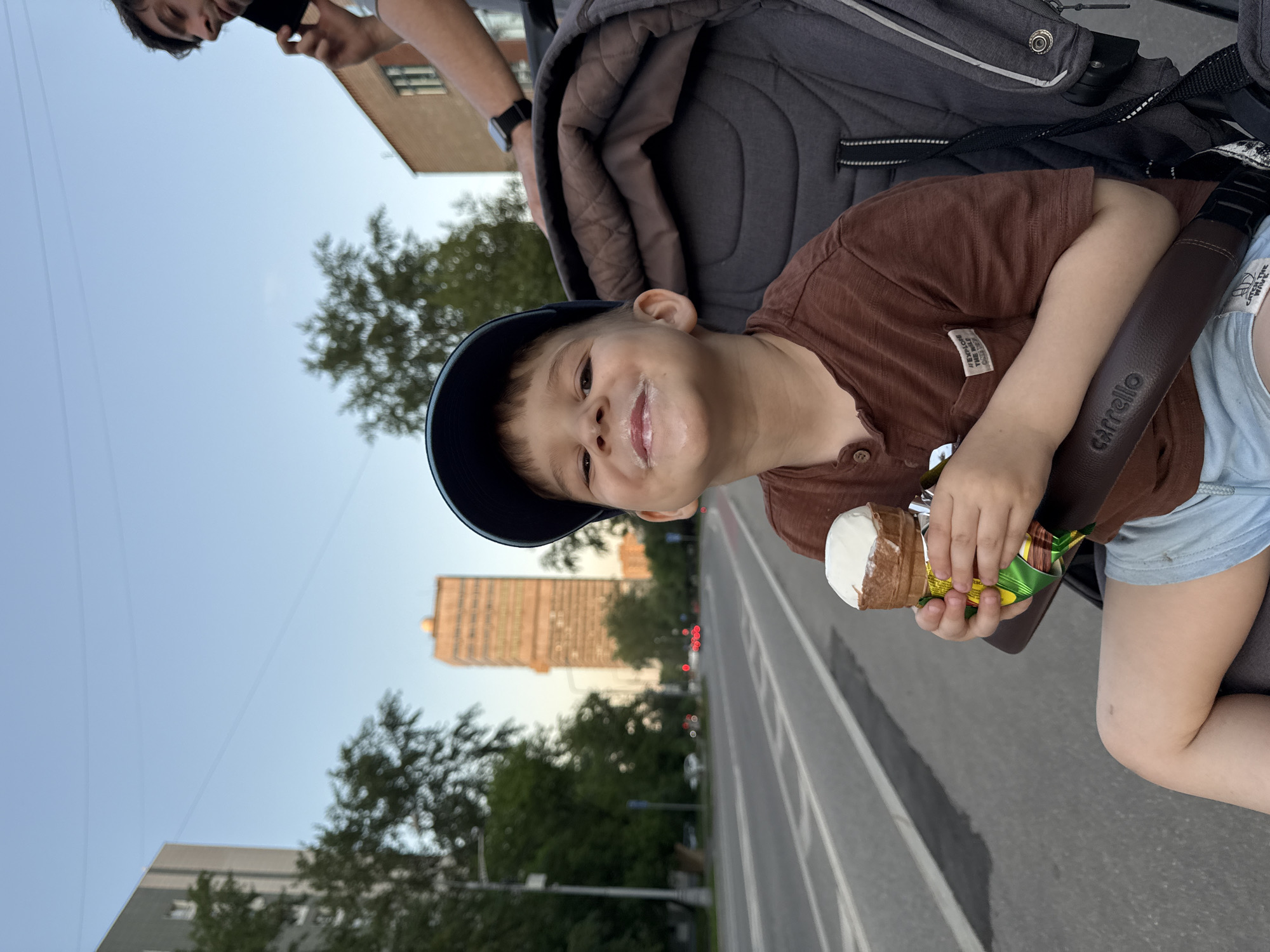 A young boy wearing a blue baseball cap, brown collared shirt, and light blue shorts is holding an ice cream cone. Some of the ice cream is on his face, specifically around his mouth. He is sitting in a stroller. A person is visible at the top left of the image, potentially pushing the stroller. In the background, there is a multi-story building, and a tree, suggesting an urban environment. The scene is set in Moscow, Russia, indicated by the "Carrello" stroller's brand, which is commonly found in Russia. The photo seems to have been taken on a road or street, as indicated by the painted lines.