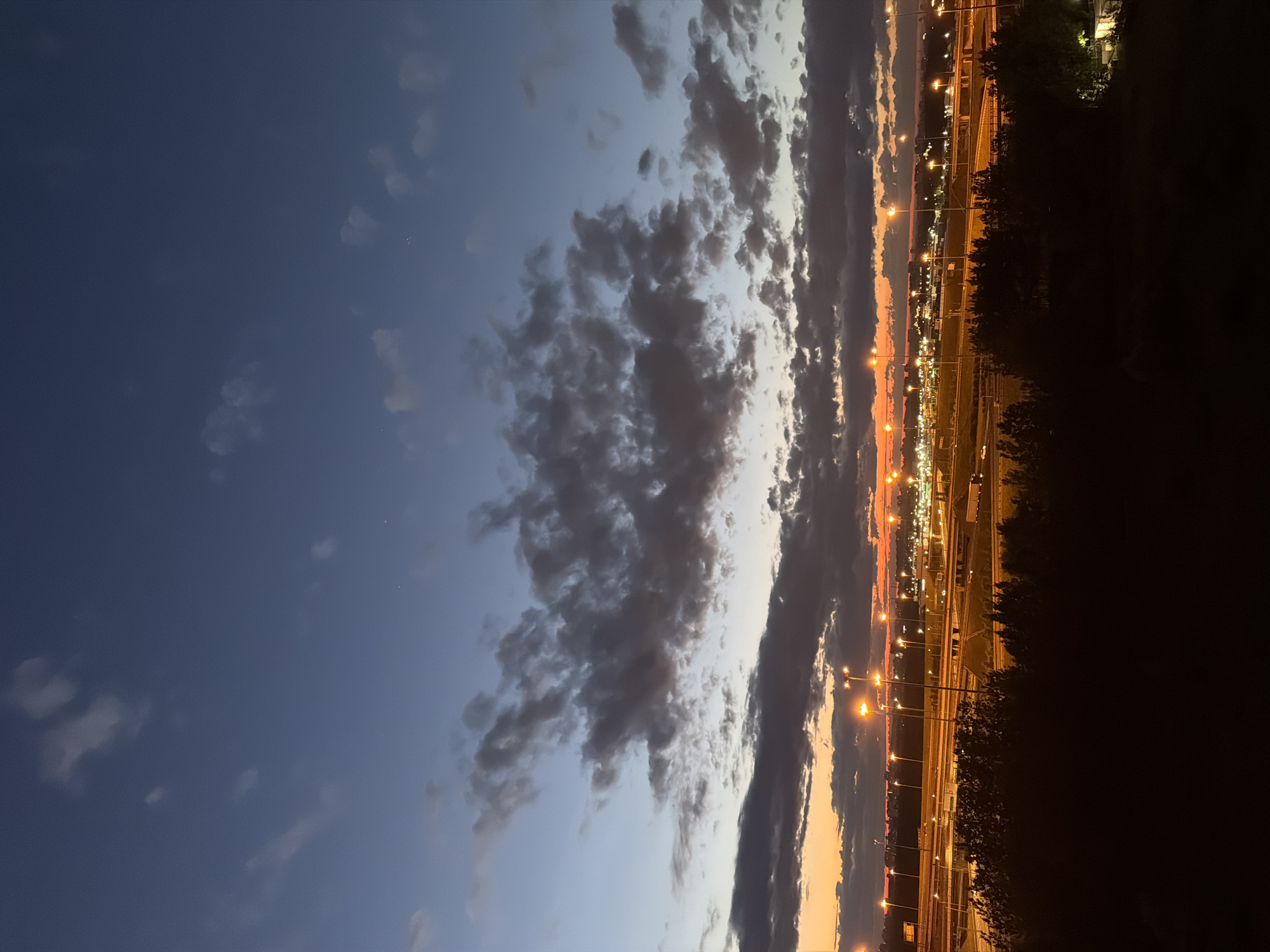 An aerial view captures a twilight scene of a highway interchange in an urban area. The sky is a gradient of deep blue at the top transitioning to a fiery orange and yellow near the horizon, punctuated by dark, textured clouds. Numerous streetlights illuminate the multiple levels of the highway and surrounding infrastructure, casting an amber glow. In the foreground, the dark silhouettes of trees and foliage obscure the immediate ground, creating a stark contrast with the brightly lit highway. The distant cityscape is visible as a hazy line of lights against the horizon.