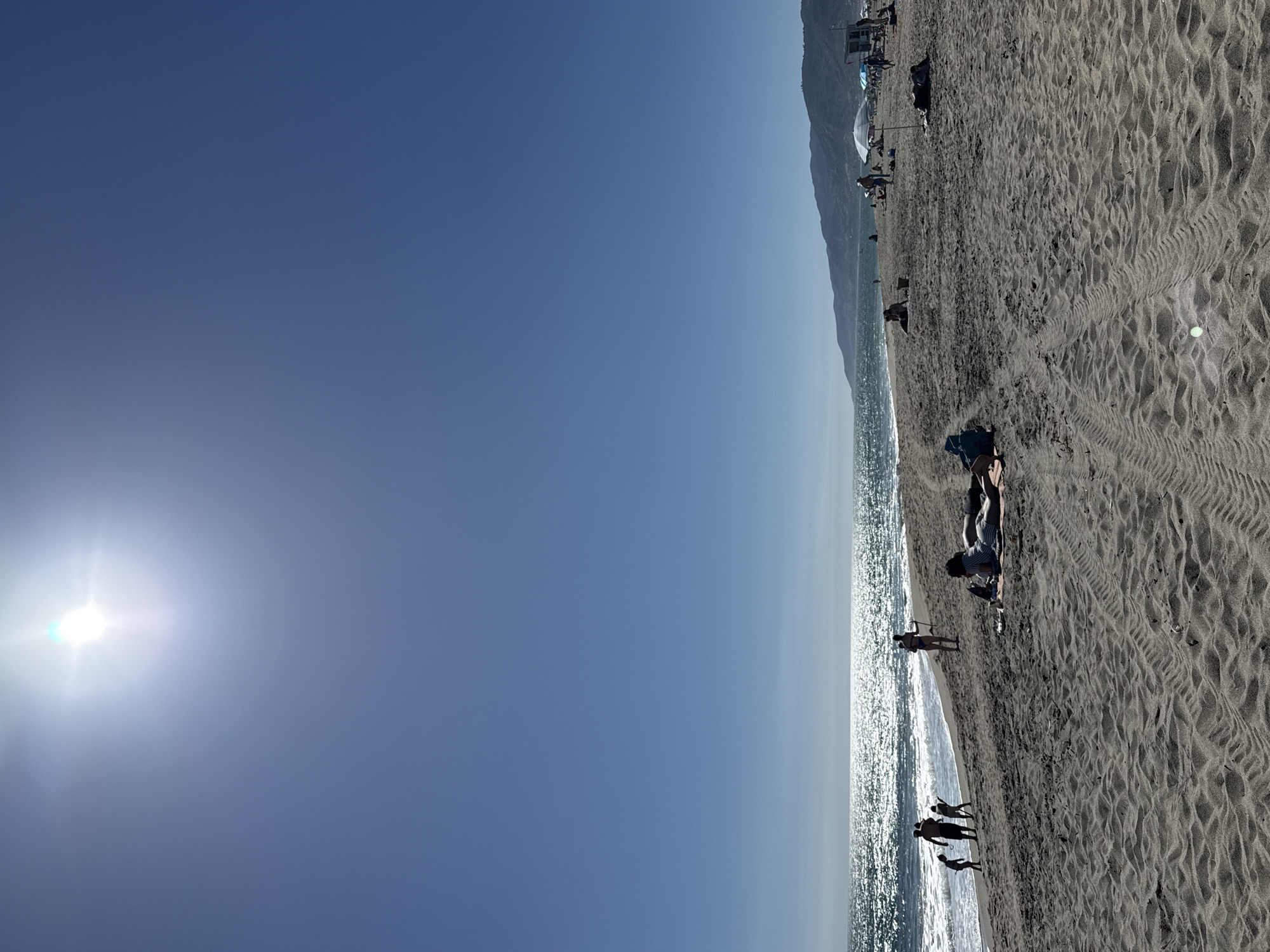 A wide, bright, sunny day is depicted on a sandy beach in Malibu, California, USA. The sun is high in a clear blue sky, casting a bright glare over the ocean. The water sparkles with sunlight, and gentle waves lap the shore. A group of people are visible in the distance, some wading in the ocean and others walking along the sand. Closer to the foreground, several individuals are relaxing on the beach, some lying on towels or blankets. Beach umbrellas and a lifeguard stand are scattered across the sand. Tire tracks are imprinted on the sand, suggesting recent vehicle activity on the beach. In the background, rolling hills form a scenic backdrop to the coastal landscape.