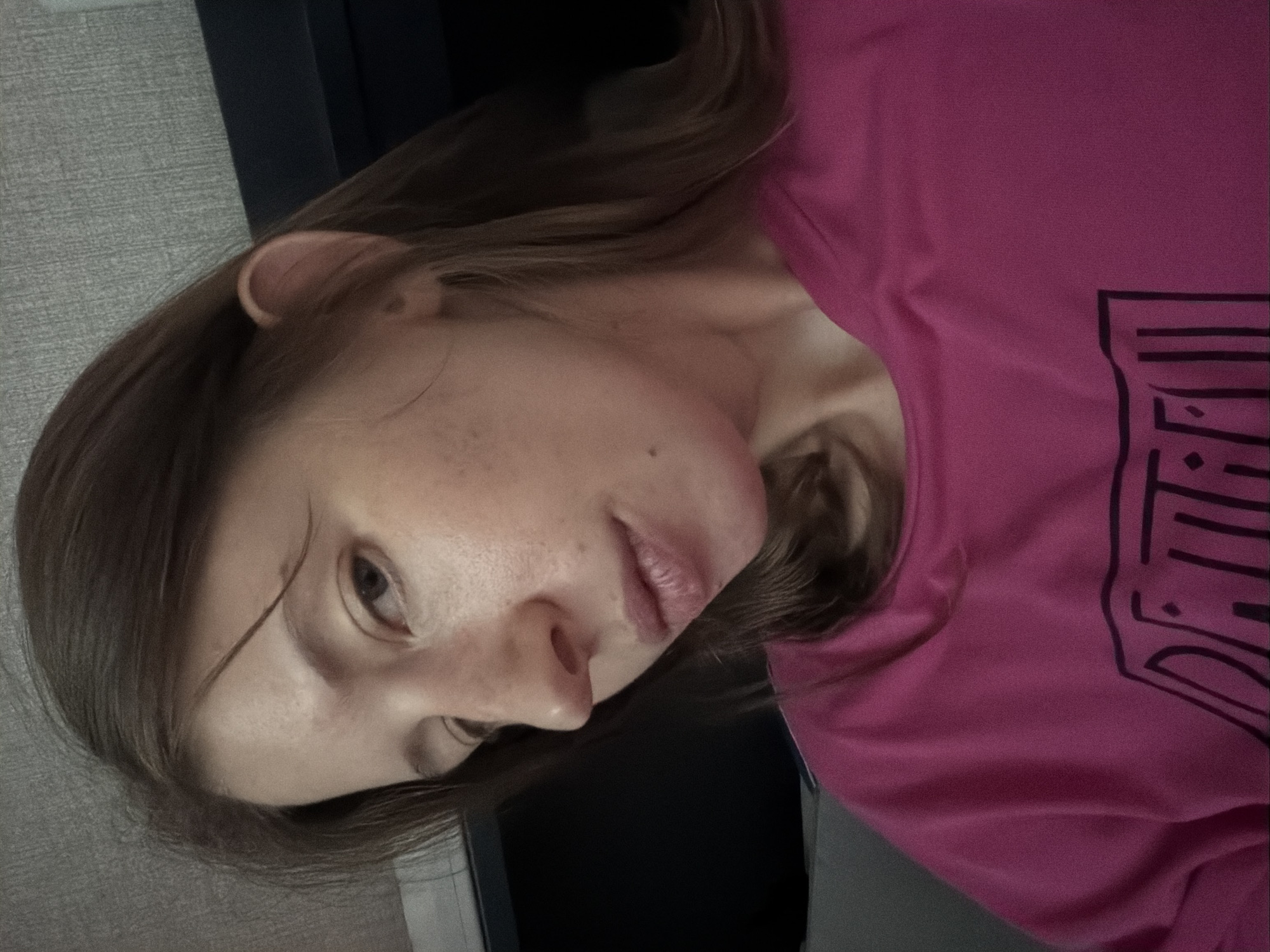 A young woman is pictured from the chest up, with her head turned to the left. She has light brown hair and fair skin with freckles. She is wearing a bright pink crewneck shirt with a black graphic logo on it. The background is out of focus and appears to be indoors, possibly a bedroom, with a gray textured wall and a dark blue headboard. There are no discernible landmarks or clues to indicate the specific location within Dongducheon-si, South Korea.
