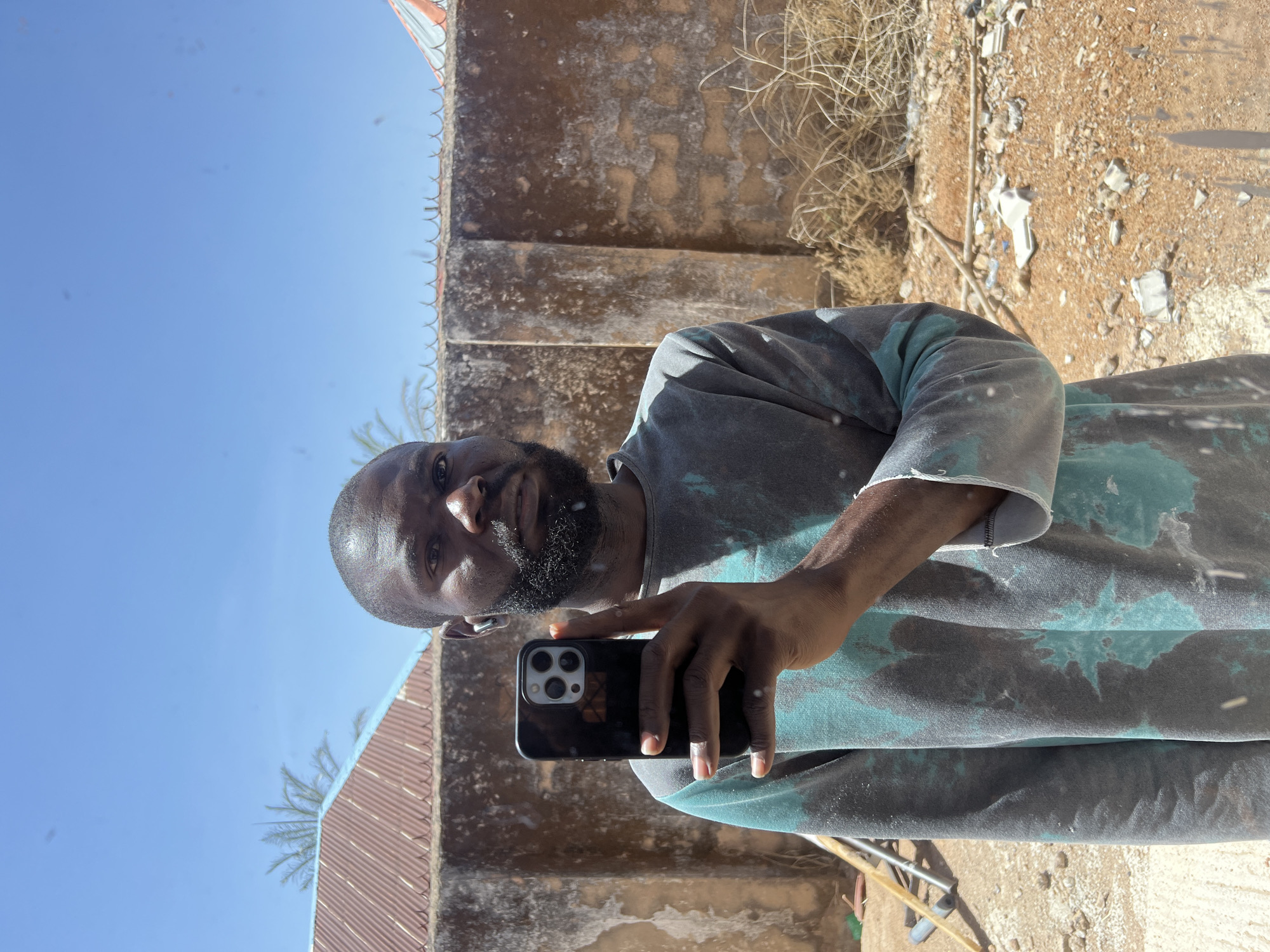 A man is seen in a close-up selfie in what appears to be Jos, Nigeria. The individual, likely in his late 30s to mid 40s, is holding a smartphone in his right hand. He has a closely-shaved head, a short, well-groomed beard, and dark skin. He is wearing a t-shirt with a teal and grey tie-dye pattern, which appears to be worn or dusted with some sort of light-colored particulate matter.
The background consists of a mottled, weathered wall with visible stains and signs of age. Part of a corrugated metal roof is visible on the left, while dry grass and debris are visible to the right. The sky is a clear blue. The ground appears to be uneven and composed of dirt and loose debris. The lighting suggests it is daytime.
