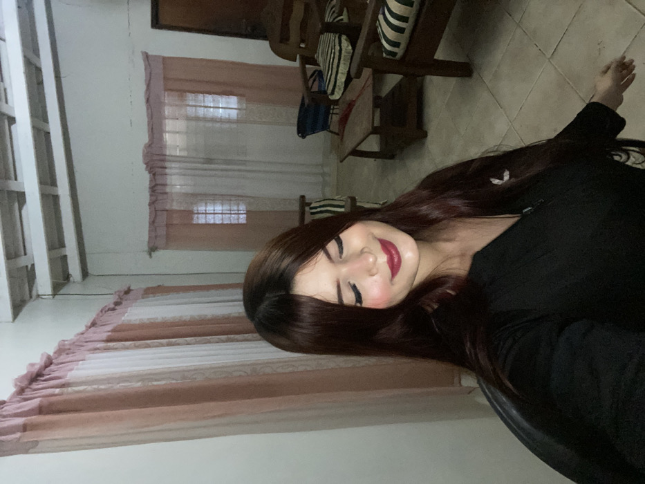 A brunette woman with fair skin and red lipstick smiles gently at the camera, wearing a black shirt. The background reveals a room with white walls, curtains, and wooden furniture, including a chair with striped cushions. Tile flooring is also visible. The image is oriented sideways.