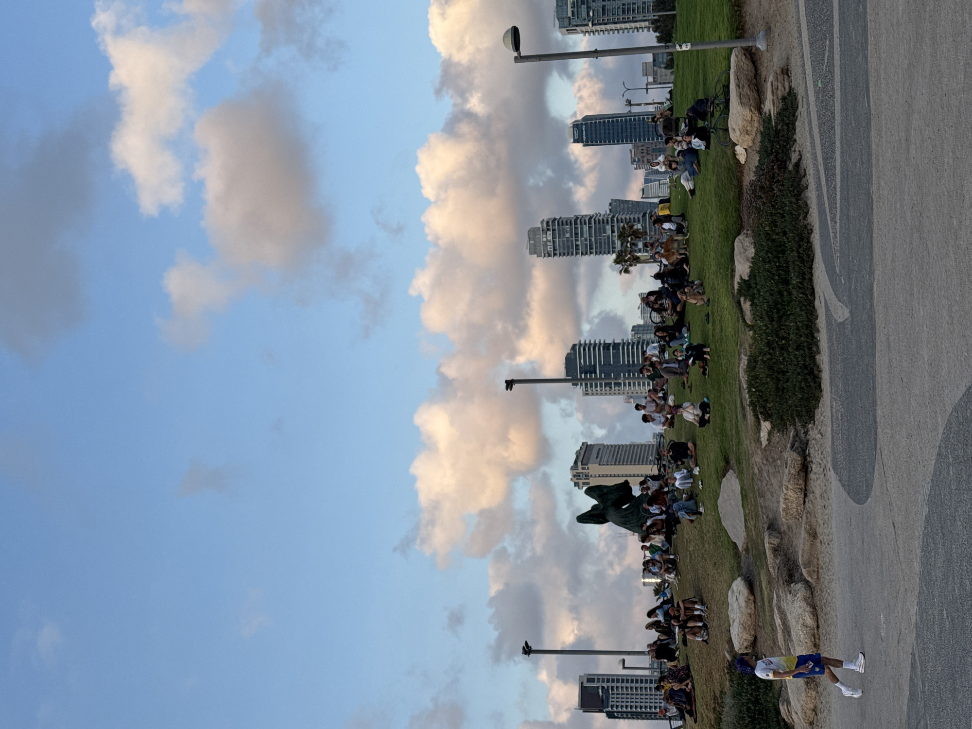 The image shows a view of the Tel Aviv coastline in Israel, with a gathering of people on a grassy slope overlooking the sea. Several high-rise buildings are visible in the background. The sky is filled with clouds. A paved pathway is on the right side of the image, with a person wearing a white shirt and blue shorts walking on it.