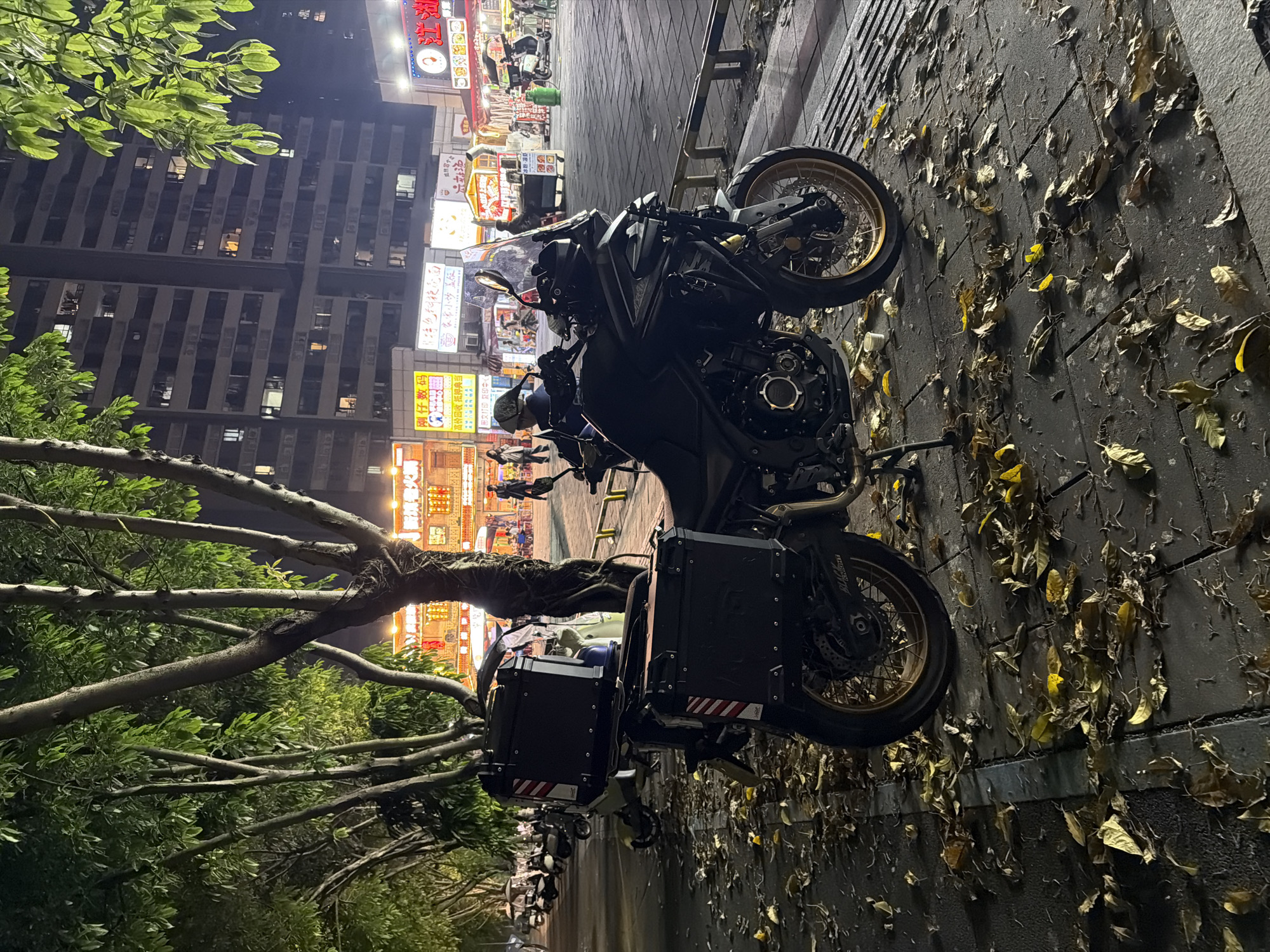 A motorcycle with black panniers is parked on a city sidewalk in Chongqing High-tech Zone, China. The sidewalk is covered with fallen leaves. In the background, a tall building is visible, along with several brightly lit storefronts with Chinese characters. A tree with branches extending over the motorcycle is on the left of the frame.