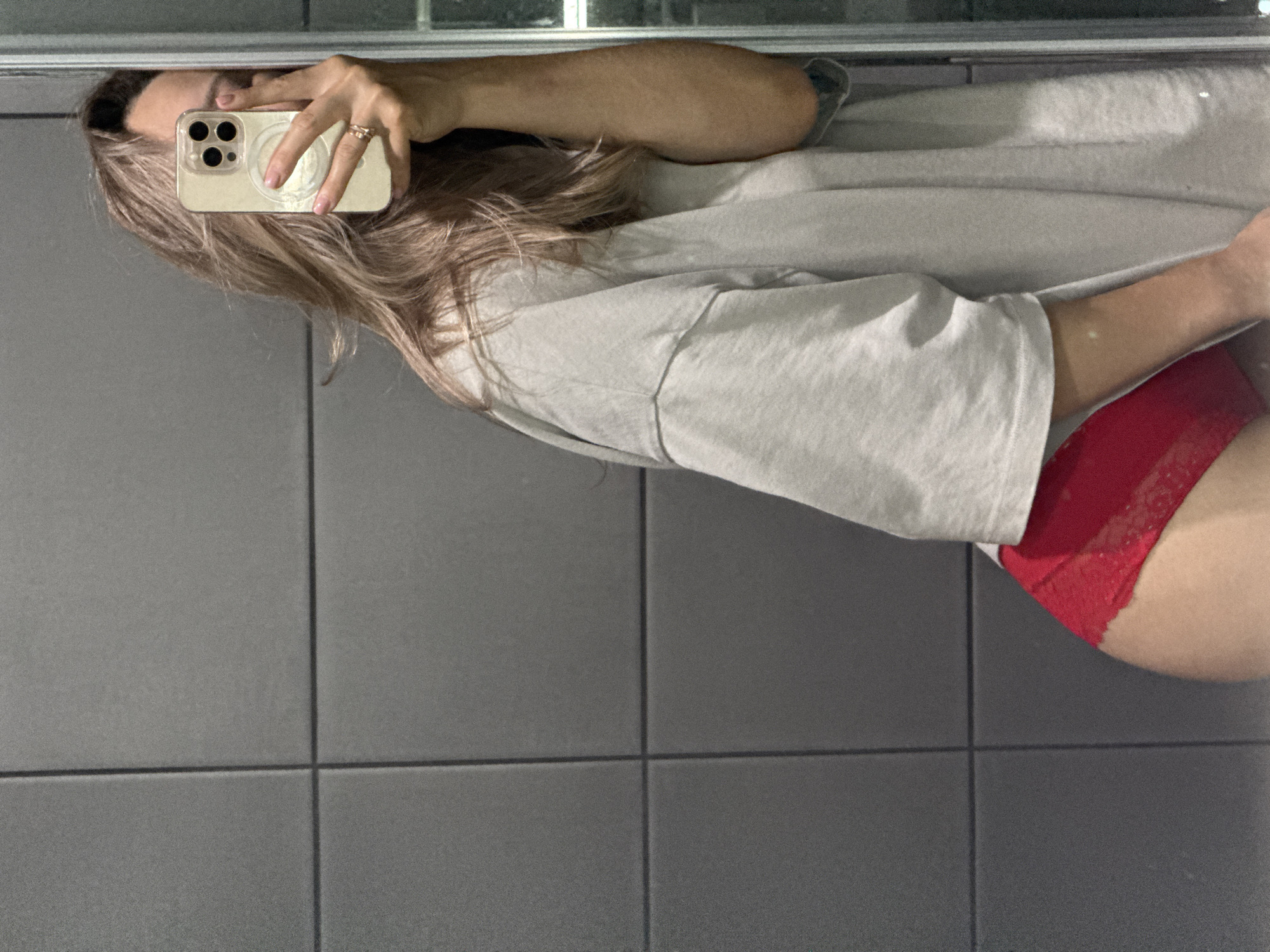 A person is taking a mirror selfie in a bathroom. They are wearing a grey oversized t-shirt and red lace underwear. The bathroom walls are tiled with dark grey rectangular tiles. The person is holding a smartphone in their right hand, obscuring their face. The image is cropped to focus on the person and their attire, with the background tiled wall prominently visible. The location is not specified in the image itself, but if it were in Chaikovsky, Russia, it would be a domestic scene in a private residence.