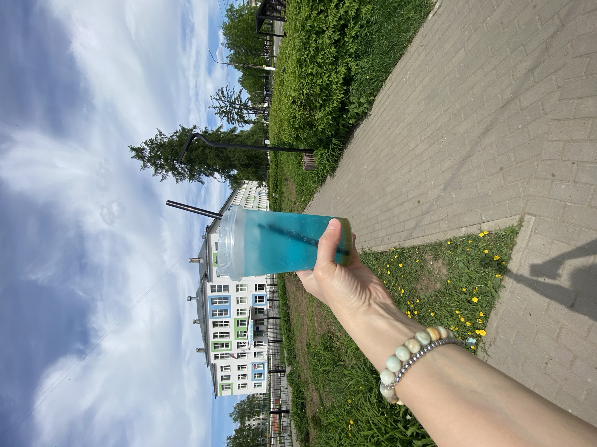 A person is holding a blue beverage in a clear plastic cup with two black straws, on a paved sidewalk next to a grassy area with yellow flowers. The person's arm is visible, adorned with a bracelet made of light green and silver beads. In the background is a multi-story building with white walls, blue and green accents, and numerous windows. The scene appears to be in Chaikovsky, Russia, as indicated by the visible architecture and overall environment. Above the building are trees and a partially cloudy sky.