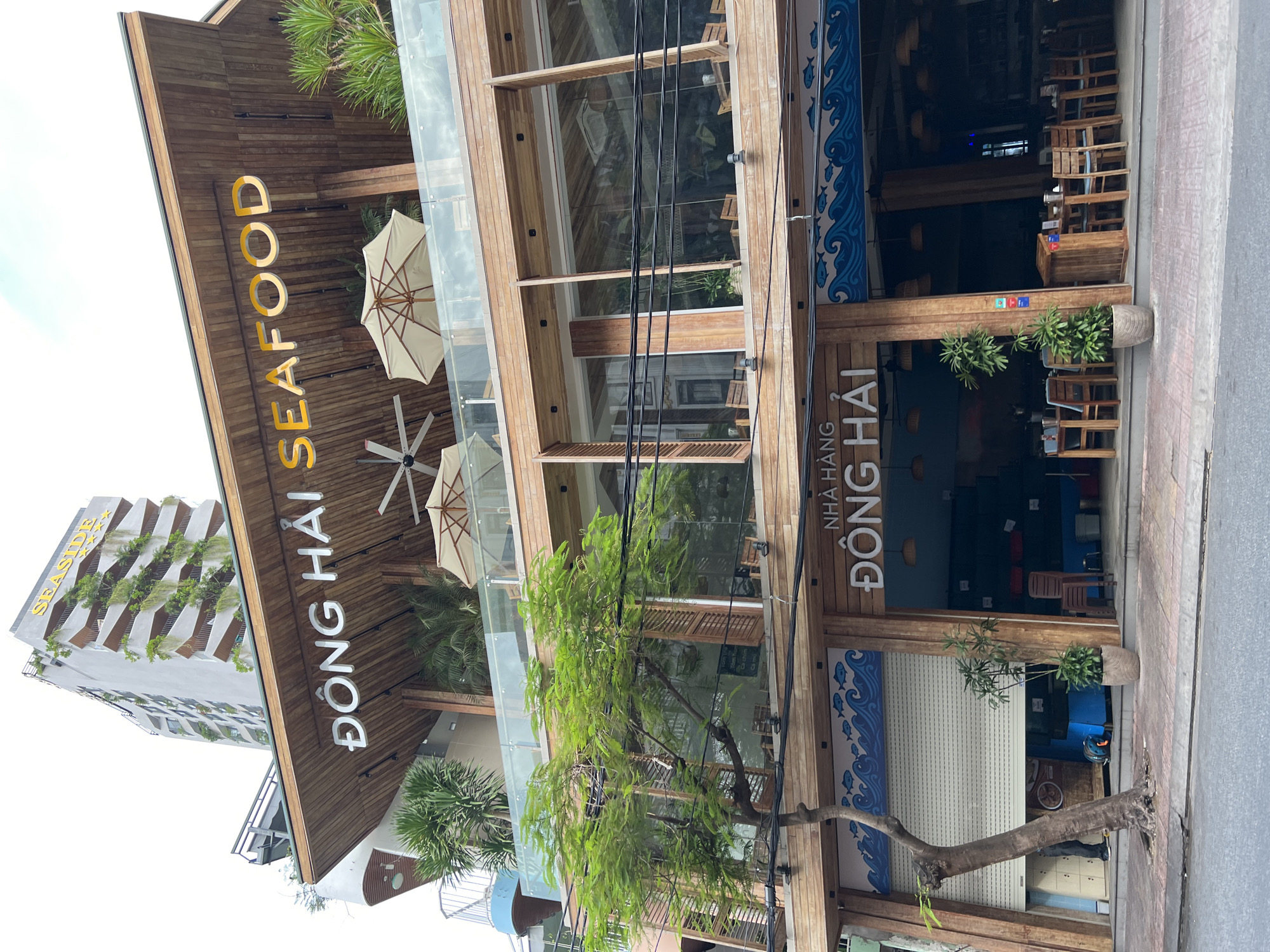 The image shows a multi-story building with a seafood restaurant called "ĐÔNG HẢI SEAFOOD" prominently displayed on its facade. The restaurant appears to be located in Nha Trang, Vietnam, as indicated by the "SEASIDE" sign on the building behind the restaurant and the architectural style. The restaurant has a modern design with wooden accents and large glass windows. There are outdoor seating areas with umbrellas and tables, and a closed storefront with a rolling metal door. A tree with green foliage stands in front of the building on the sidewalk. The street in front of the restaurant is visible, with a sidewalk and road. The sky is overcast.
