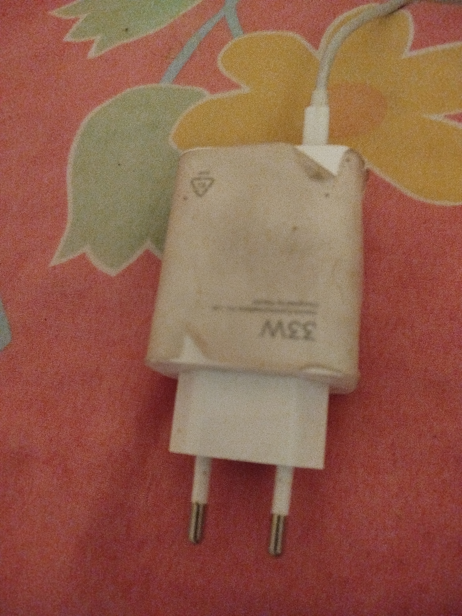 The image shows a close-up, slightly blurry, overhead view of a white phone charger with visible "33W" markings on its side. The charger is plugged into a wall outlet, with its prongs extended downwards. The background is a patterned fabric, possibly a bedsheet or tablecloth, with a red base color and abstract floral designs in green and yellow. There are no people visible in the image, nor any indication of a specific location in Lassan, Pakistan. The scene appears to be an indoor setting.