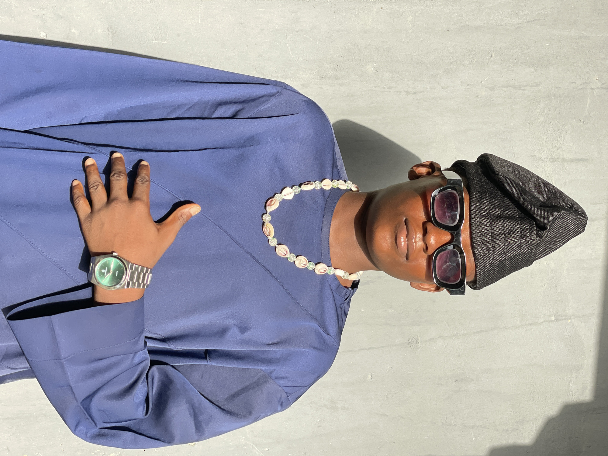 A portrait-style image, taken in Badagry, Nigeria, features a person of color looking directly at the camera. The subject is dressed in a deep navy blue long-sleeved garment, possibly a tunic or kaftan, which appears to be made of a smooth fabric. Their right hand, adorned with a silver-toned watch featuring a circular green face, rests gently on the chest area of the garment.
The person wears several distinct accessories: a black, textured, possibly woven cap or headwear is perched on their head, and stylish dark-framed sunglasses with reddish-purple tinted lenses cover their eyes. Around their neck is a necklace crafted from light-colored, irregular shell-like beads interspersed with smaller, clear or light green beads. The subject has a neutral, composed expression with slightly parted lips.
The lighting is bright and direct, suggesting a sunny outdoor setting during the day, likely around midday or early afternoon, as evidenced by the sharp shadows cast. The background is a plain, light gray, textured surface, possibly concrete. The photograph is oriented vertically, showing the subject from the chest up. There is no visible text in the image.
