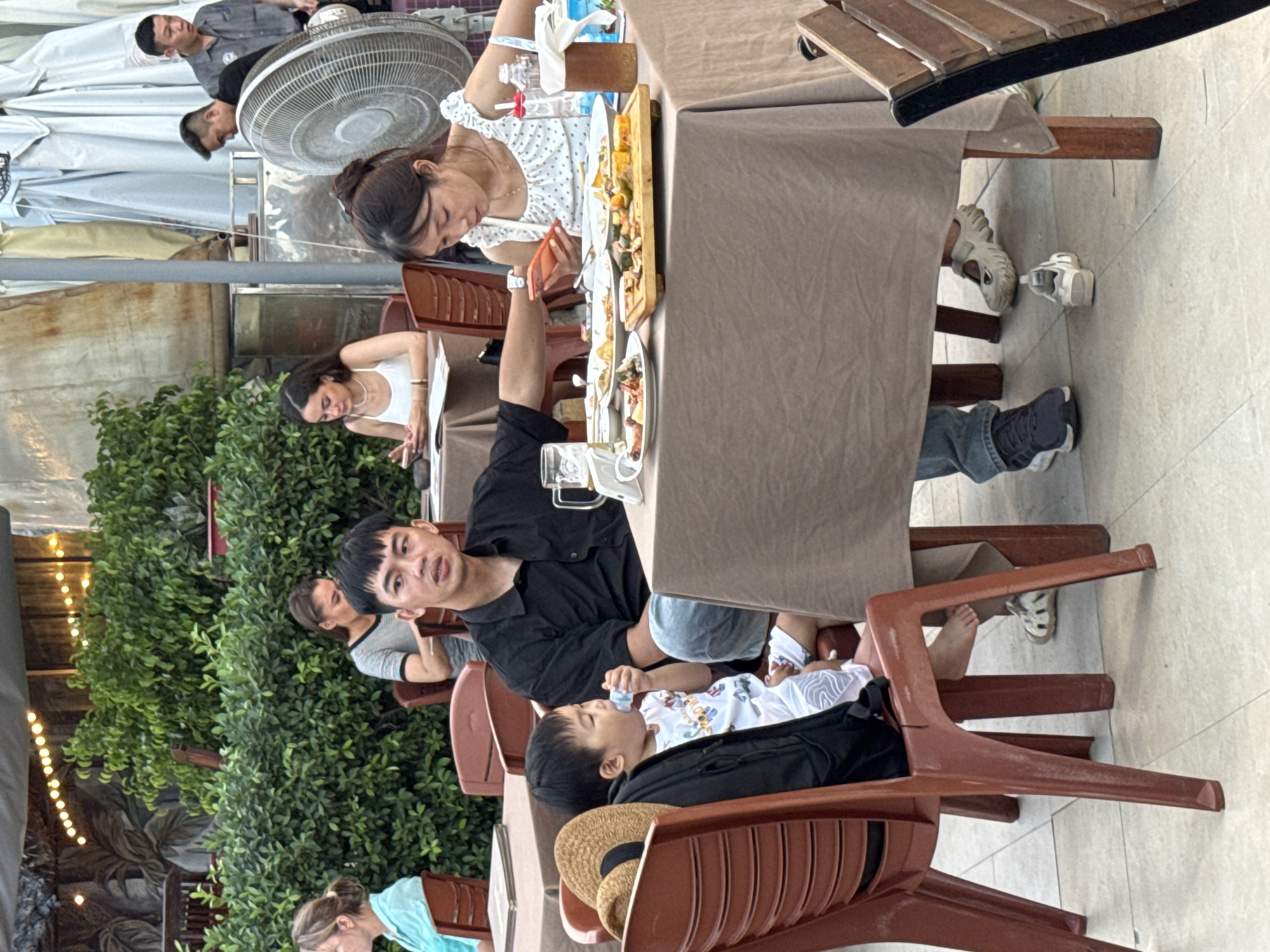 A group of people are dining at an outdoor restaurant with tables covered in brown tablecloths. In the foreground, a young child wearing a white shirt and shorts sits in a red plastic chair, holding a bottle to their mouth. Next to the child, a man in a black collared shirt and blue jeans is seated. Across from them, a woman in a white polka-dot top looks down at her orange cell phone. In the background, other diners and staff are visible. The setting appears to be a casual eatery in Nha Trang, Vietnam, with lush green bushes and outdoor lighting contributing to the ambiance. A large industrial fan is also present, suggesting a warm climate.
