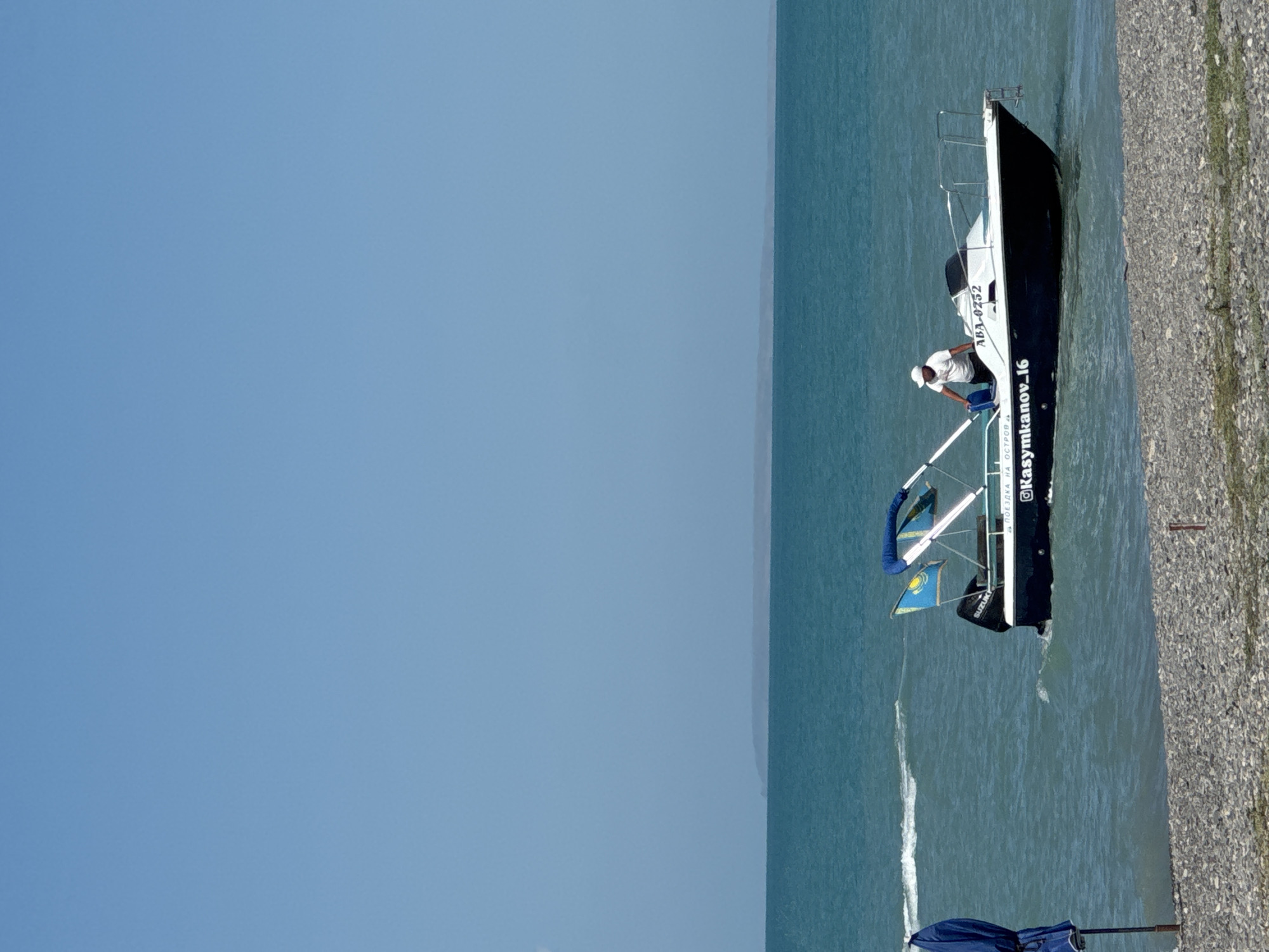 A motorboat with a white and blue canopy is on the water near a pebble beach. A man wearing a white t-shirt and a white cap is standing in the boat. There are two flags on the boat, one of which appears to be the flag of Kazakhstan. The boat has the registration number "ABA-0752" and text that reads "@Kasymkanov_16" and "ПОЕЗДКА НА ОСТРОВ". A blue beach umbrella is visible on the left side of the image. The water is a turquoise color, and there is a distant island or landmass on the horizon under a clear blue sky. The scene is set in the city of Zharbulak, Kazakhstan.