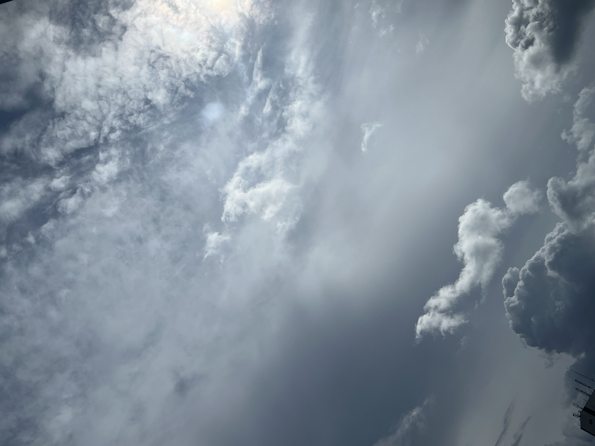 The image is a low-angle shot of a cloudy sky, taken from a city, as indicated by the building structure visible at the bottom left. The sky is filled with various cloud formations, ranging from thin, wispy cirrus clouds to larger, more defined cumulus clouds. Some parts of the sky are brightly lit, suggesting the sun is either present or just behind the clouds, while other areas are darker and appear more ominous, possibly indicating approaching weather changes. The overall color palette is dominated by shades of gray and white, with a hint of blue in some clearer patches. The composition is largely abstract, focusing on the dynamic textures and patterns of the clouds. There are no people or specific actions depicted in the image. The image does not contain any information to suggest the location is Warsaw, Poland.