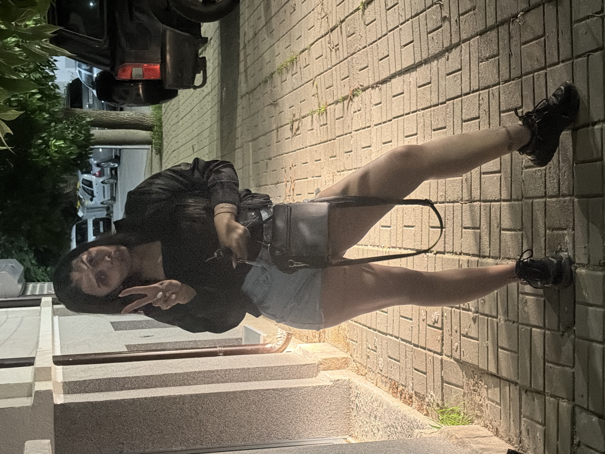 An outdoor, nighttime full-body shot features a young woman posing on a paved walkway next to a set of concrete stairs in Shumen, Bulgaria. The woman, with dark hair, is wearing a black long-sleeved top or jacket, light blue denim shorts, and black sneakers. She holds a black handbag over her left shoulder and is making a peace sign with her left hand, which has white-painted nails. Her right arm is slightly bent at the elbow. She has a faint smile on her face.
The setting appears to be an urban or residential area. The ground she stands on is paved with rectangular block-style stones, with some small weeds growing in the cracks. To her left are concrete stairs with a dark railing. In the background, out of focus, are parked cars, including a dark vehicle with a red taillight visible at the top of the frame. Lush green foliage and trees are also visible further back. The scene is dimly lit by artificial light sources, casting some shadows and highlighting the woman and parts of the pavement. No visible text is present. The overall mood is casual and relaxed.