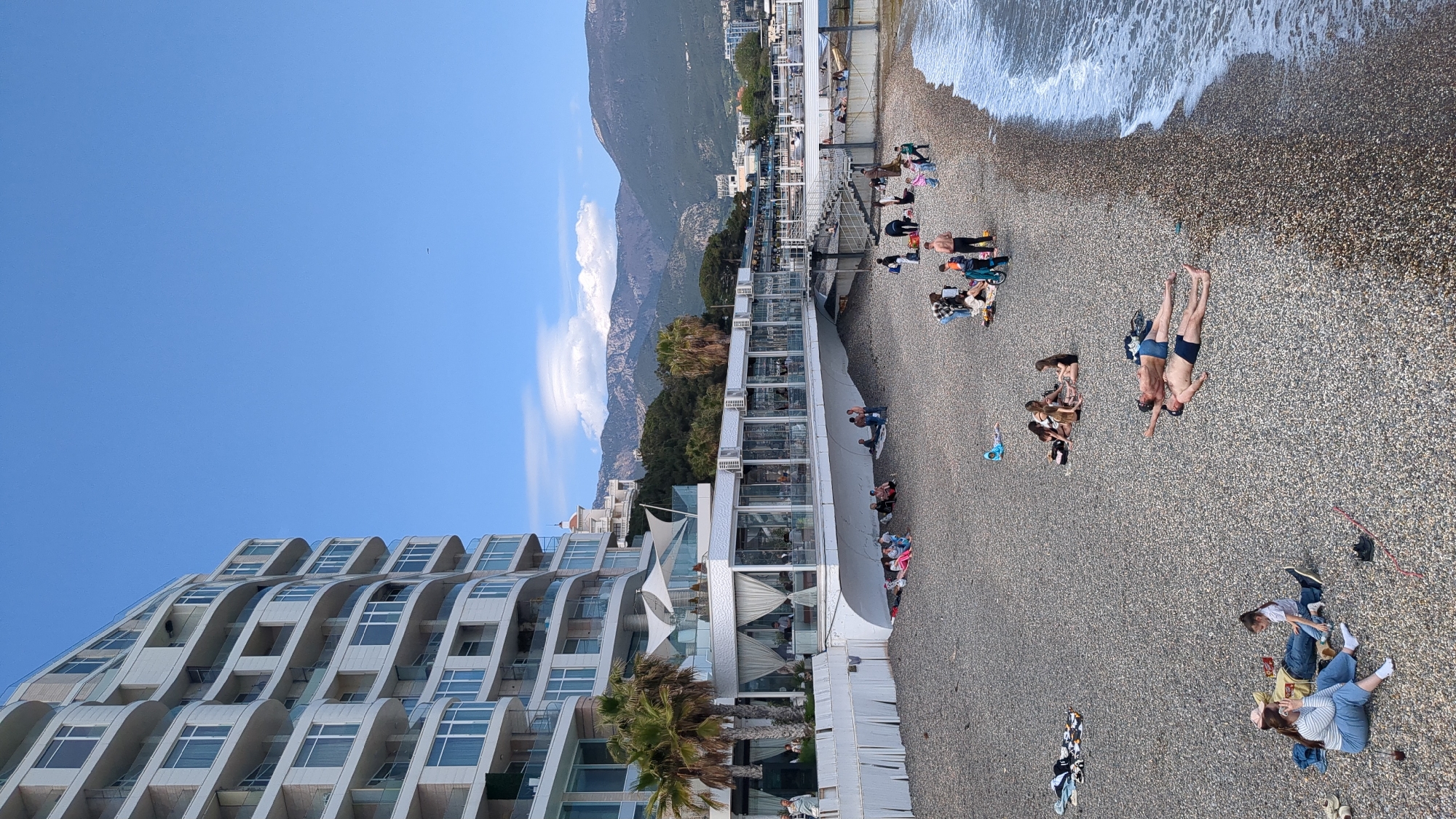The image depicts a beach scene in город Ялта, Ukraine, featuring a pebble beach, the Black Sea, and surrounding architecture. People are engaging in various activities on the beach, including sunbathing, lounging, and picnicking. Two individuals are lying on the pebbles, seemingly sunbathing. The shoreline is visible with gentle waves lapping at the shore.
A multi-story hotel with curved balconies is prominently featured to the left of the frame. Adjacent to the hotel is a white structure, possibly a restaurant or beach club, with open-air sections and shaded areas. Trees and vegetation are interspersed between the buildings and the beach. In the background, a mountain range rises. The sky is mostly clear blue with some scattered white clouds.