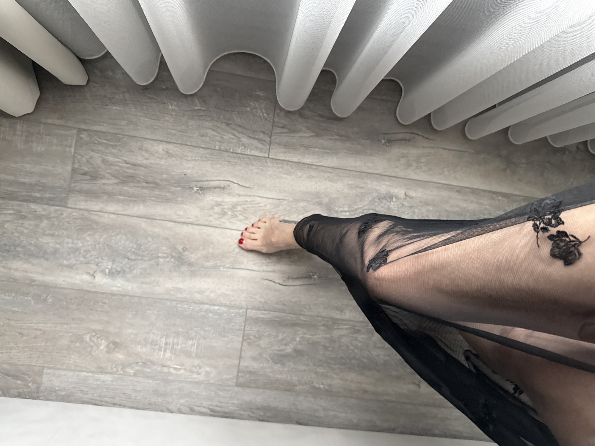 A low-angle, eye-level shot depicts a woman's leg clad in a black, sheer garment, likely lingerie or a dress, adorned with black floral appliqués, resting on a gray wood-look floor. The woman's foot is visible with red toenails. The backdrop consists of white curtains. The location is assumed to be an interior space in the city of Khimki, Russia.