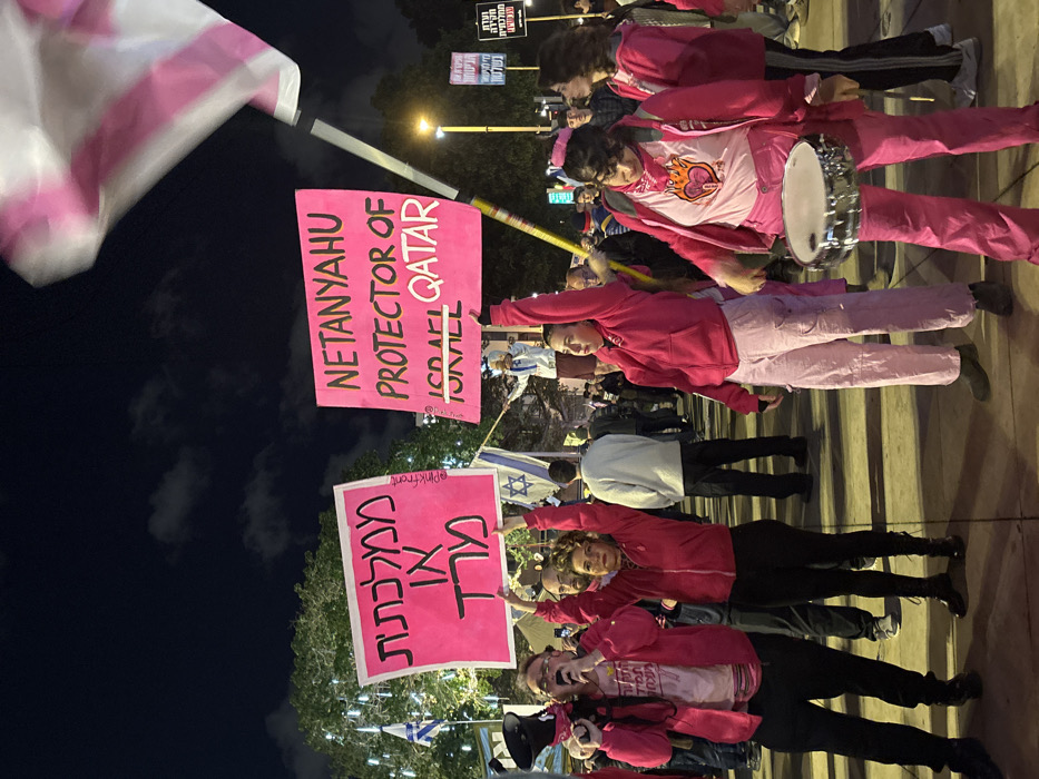 A nighttime protest shows people holding pink signs. One sign reads "Netanyahu Protector of Qatar." Another sign with Hebrew text is also visible. Some protesters wear pink clothing, and Israeli flags are interspersed throughout the crowd. A person plays a small drum and the atmosphere suggests a political demonstration or rally.