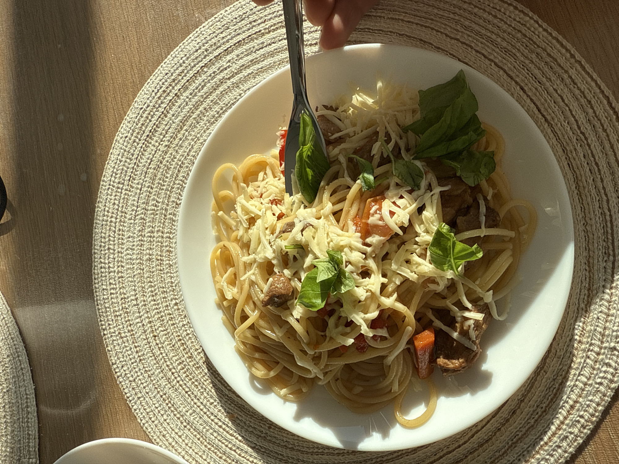A top-down, sunlit shot shows a plate of spaghetti with meat sauce, shredded cheese, and basil leaves being eaten with a fork. The plate rests on a round, woven placemat. The table is partially visible on the left, with a glimpse of another white dish. The location is unknown and there is no evidence to suggest this photo was taken in Perm, Russia.