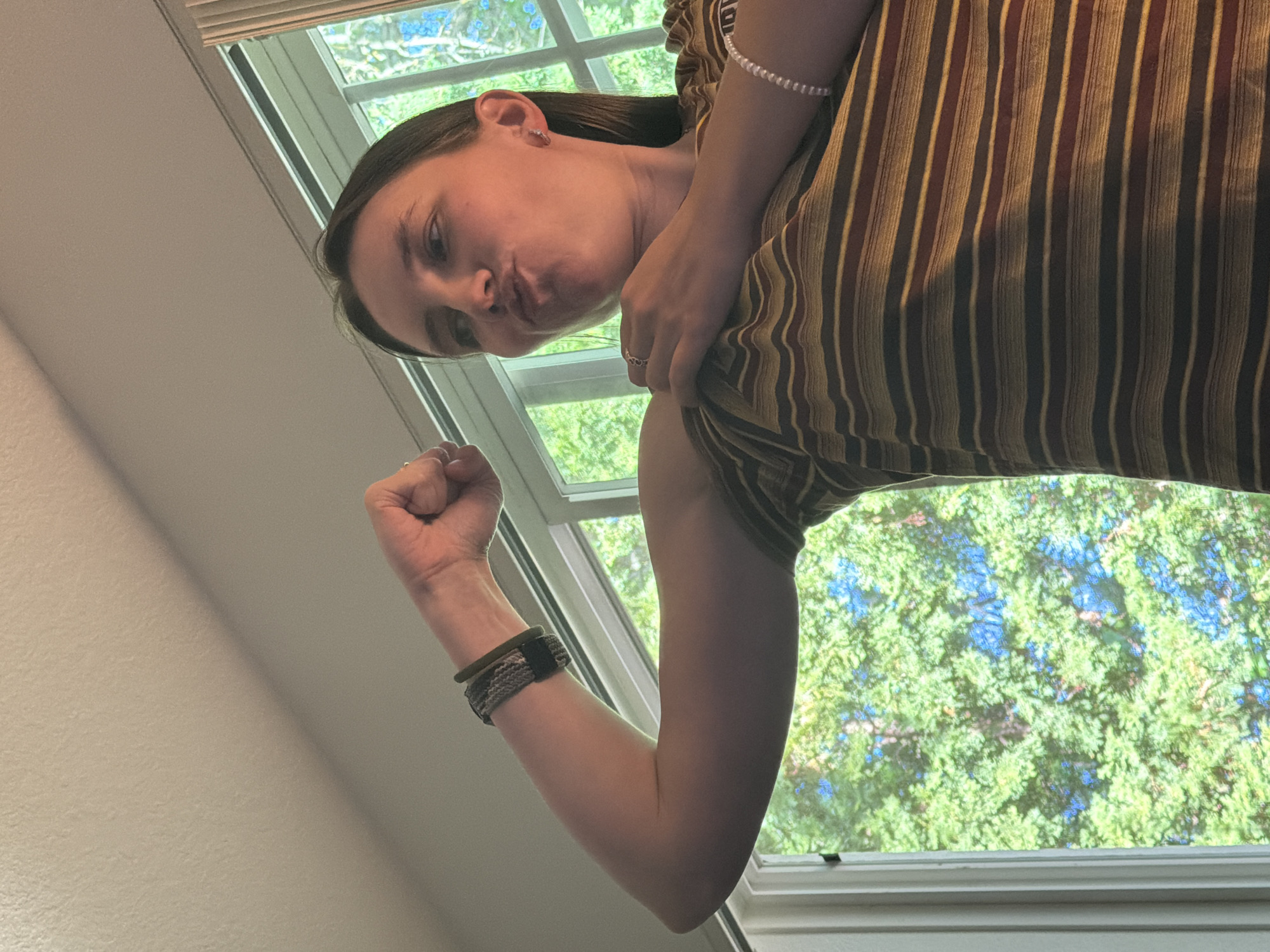 A young woman is shown from the chest up, flexing her bicep in front of a window. She has dark hair and is wearing a striped t-shirt with horizontal bands of yellow, dark red, and brown. She also has bracelets on both wrists, one appearing to be a woven band on her left arm and a string of white beads on her right. Her facial expression is one of playful exertion, with her lips pursed. Through the window, green trees with patches of blue sky are visible. The lighting suggests it is daytime, likely indoors near a window. The specific location in Portland, United States of America, cannot be determined from the visual information provided.