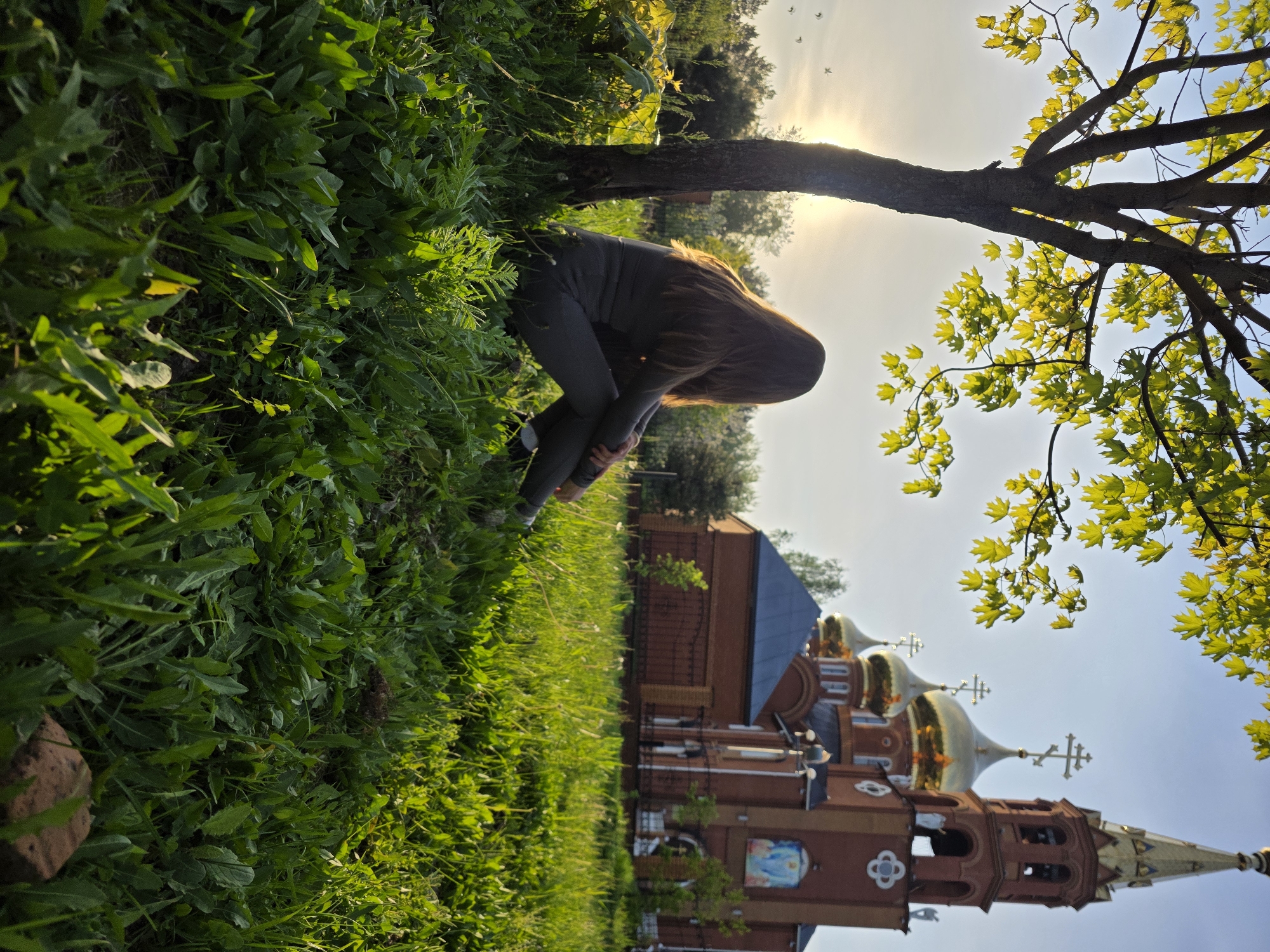 The image shows a woman with long brown hair crouching near a tree, with the Russian Orthodox Church of the Holy Great Martyr George the Victorious in the background. The church is located in Chaikovsky, Russia, visible with its red brick walls, blue roof, gold domes, and crosses. Lush green grass and foliage surround the area. The sun is shining through the trees, creating a bright and airy atmosphere.