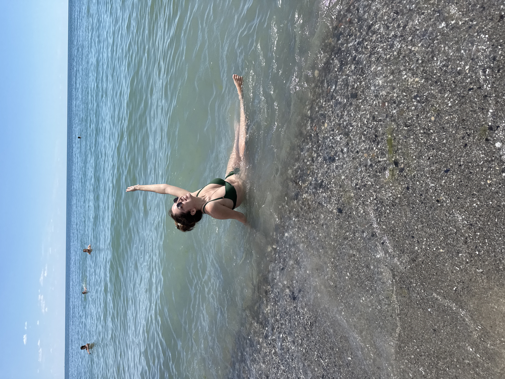 A young woman in a green bikini floats on her back in the shallow water at the edge of a pebbly beach. Her eyes are closed and she appears to be relaxed. Her right arm is extended upwards, her left hand is resting on her stomach. The water is clear and calm, with small waves lapping at the shore. In the background, several other people are swimming and enjoying the water. The sun is shining, suggesting it is a warm, clear day. The scene appears to be a typical summer day at the beach. The location is Odesa, Ukraine. There is no visible text in the image.