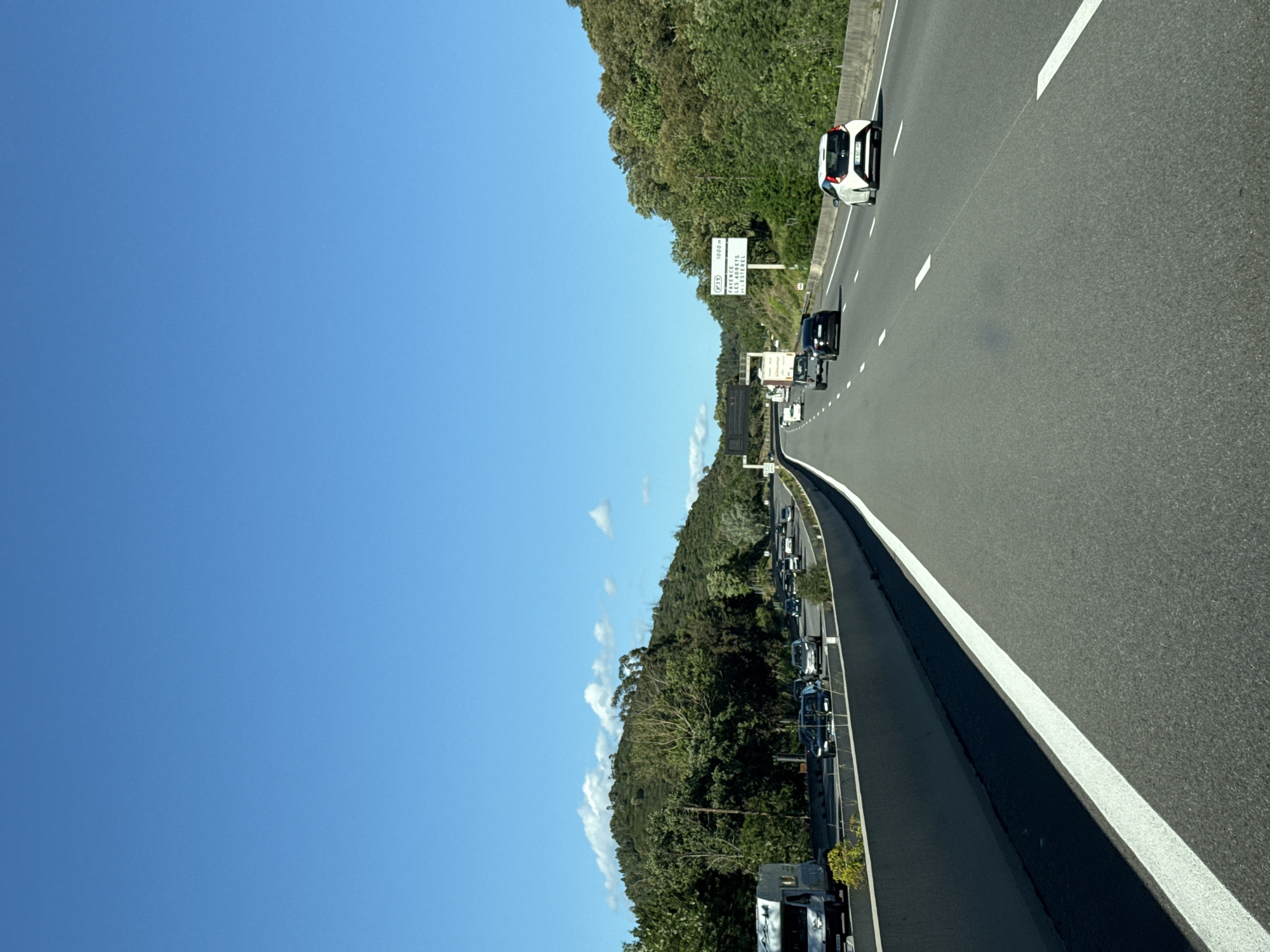 The image depicts a highway scene with a view of multiple vehicles traveling on the road. A clear blue sky occupies a significant portion of the image. The highway is flanked by green foliage, suggesting a rural or semi-rural setting. There is signage visible along the roadside. The cars are spaced intermittently, but traffic appears to be flowing. The highway appears to be located near Montauroux, France.