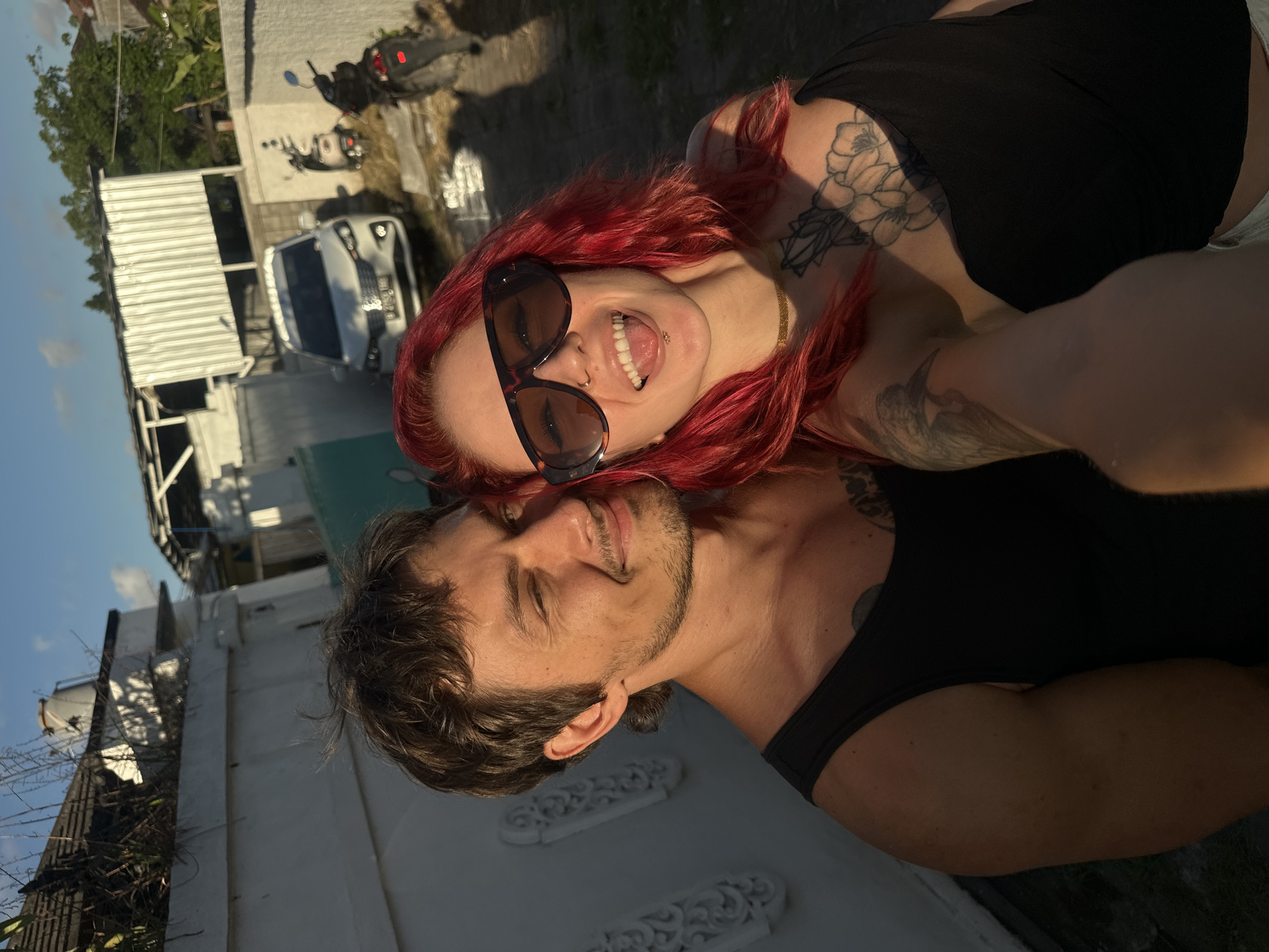 A selfie shows a fair-skinned man and woman outside. The man is tan with short brown hair, a mustache, and a goatee. He is wearing a black tank top and is smiling. The woman has red hair, fair skin, and is wearing sunglasses and a black top. She has tattoos on her neck and arm. She is sticking her tongue out and smiling. The background includes a white wall, a white van, two motorcycles, and buildings. The photo was taken in North Kuta, Indonesia.