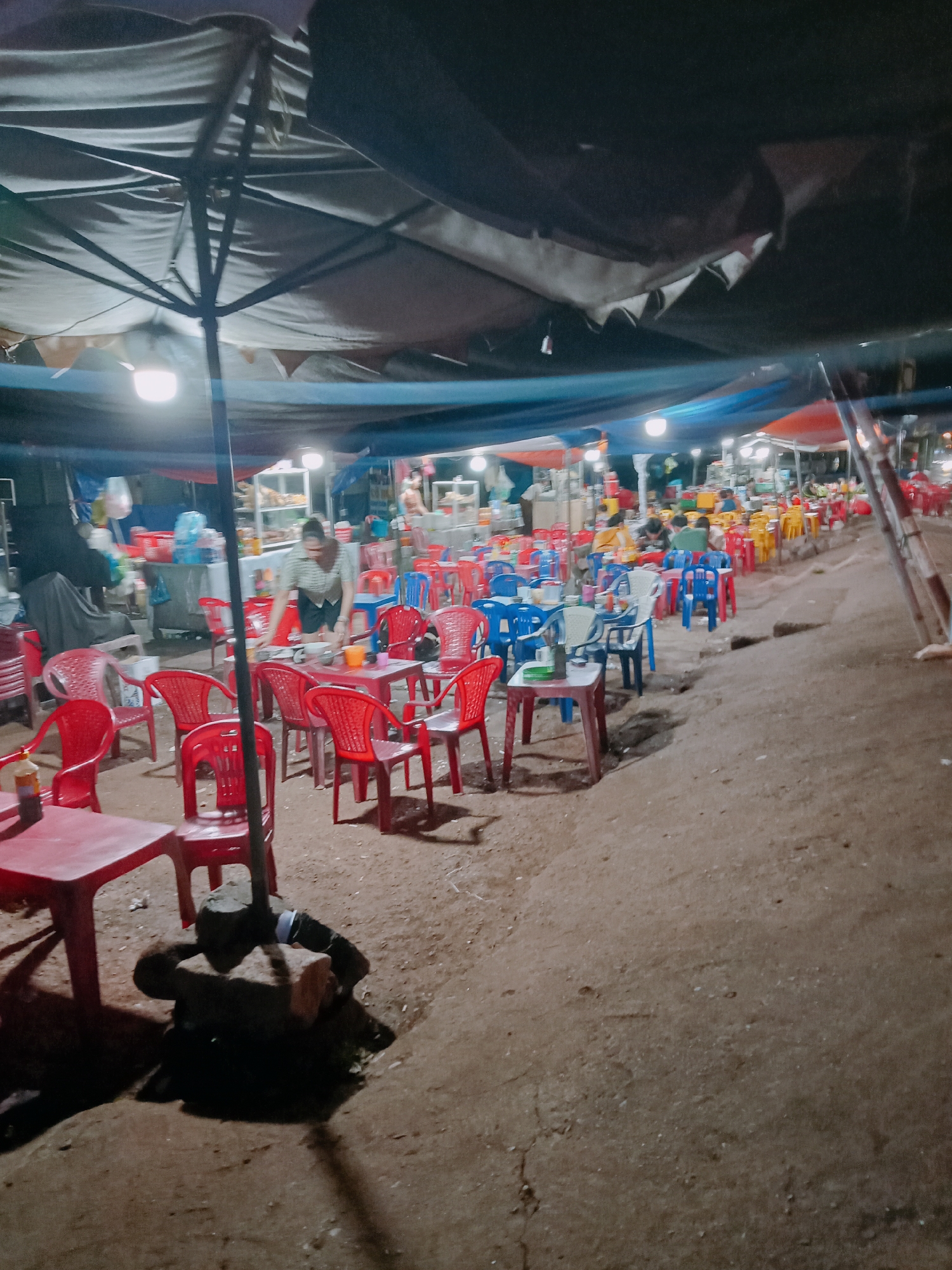 Captured in Ea Drăng, Vietnam, the image shows an outdoor dining area. A tent with visible supports covers a space filled with red, blue, and yellow plastic tables and chairs. People sit at some tables, and one person stands serving food or cleaning up. The ground is dirt, and a dark object sits at the base of a table in the foreground. String lights provide illumination.