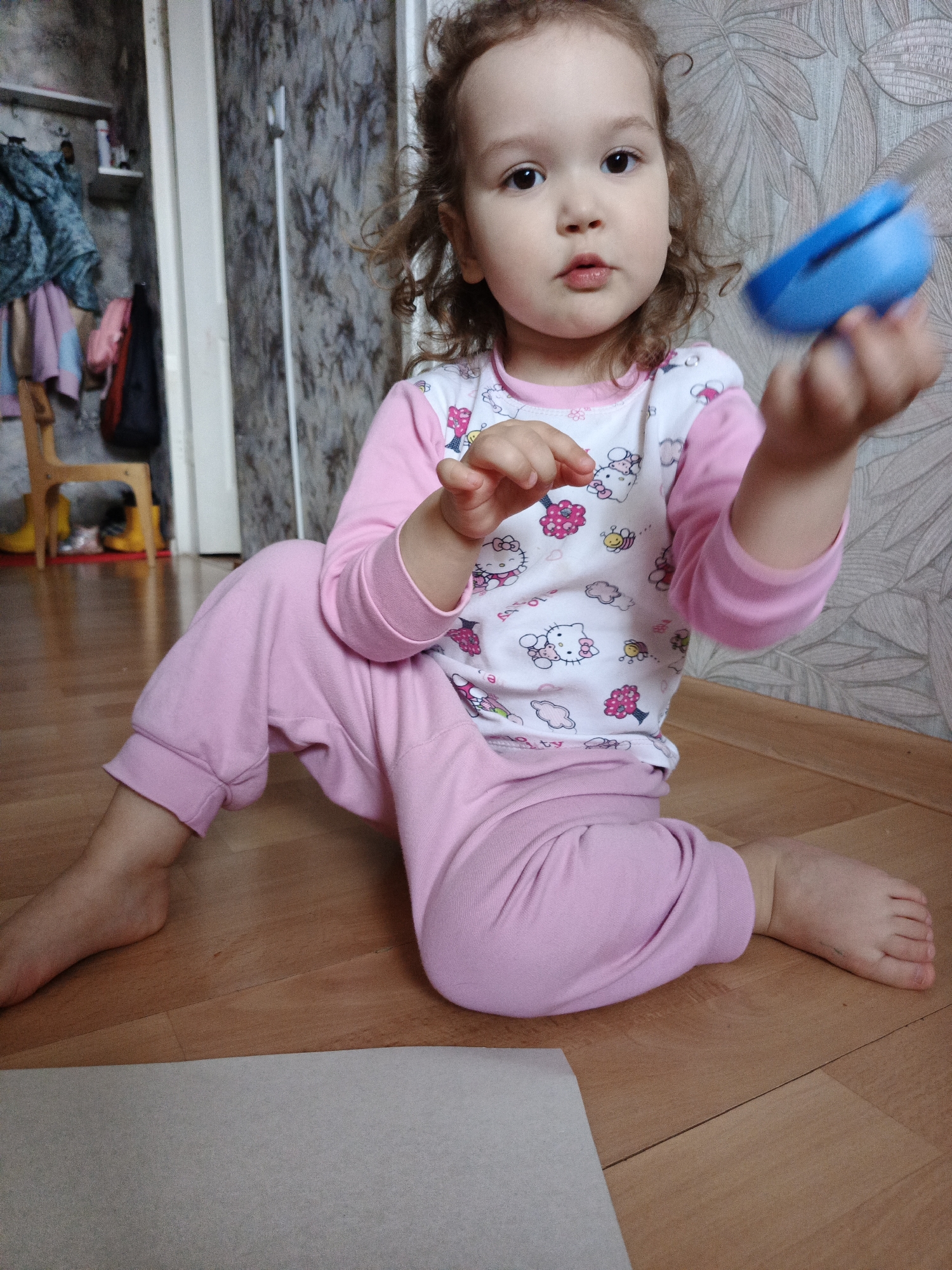 A young girl is sitting on a wooden floor in an indoor environment. She is wearing pink pajamas with a white, long-sleeved top printed with Hello Kitty and floral designs. In her right hand, she is holding a blue object. Her left hand is raised with her fingers slightly bent. She has curly brown hair and is looking towards the viewer.
Behind her is a patterned wallpaper. To the left, there is a partial view of an open closet with clothing hanging inside and a small wooden chair nearby. A white rectangular object is on the floor in the foreground. The location is a residence.