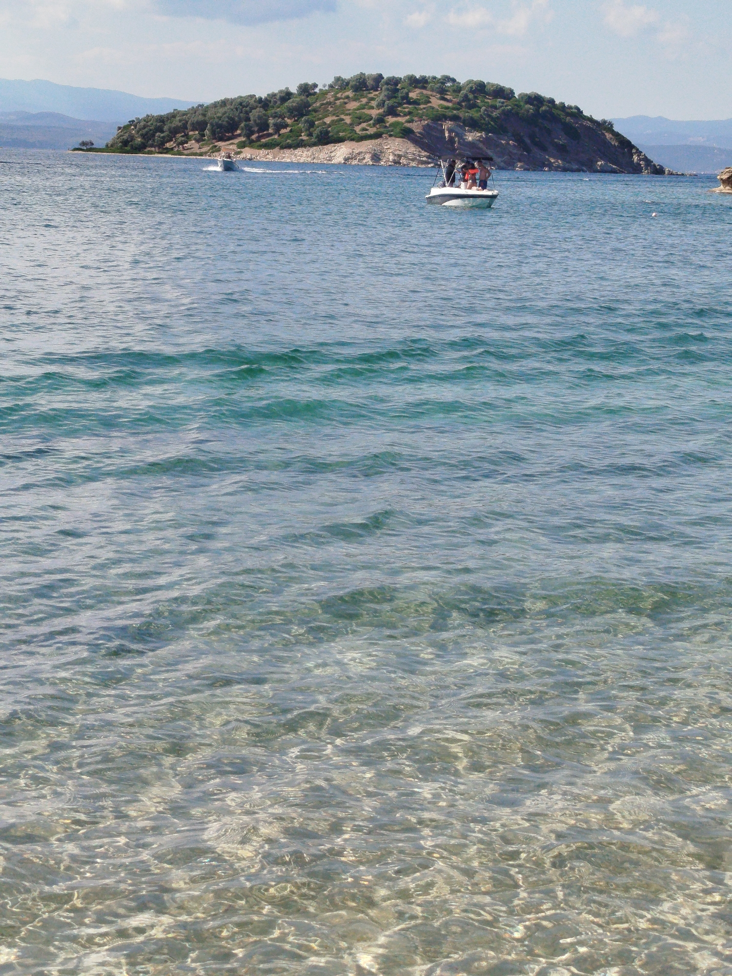 A vertical, eye-level shot captures a body of water under a partially cloudy sky. In the distance, a small island is densely covered with green foliage and sparse trees, with rocky terrain visible. A boat with several people aboard cruises towards the right side of the frame, leaving a wake behind it. Another, smaller boat is further to the left, also moving away from the viewer. The water in the foreground is clear, revealing the sandy seabed below, with gentle ripples creating patterns on the surface. The scene is likely situated in the Sithonia Municipal Unit, Greece, given the characteristic landscape and clear waters.