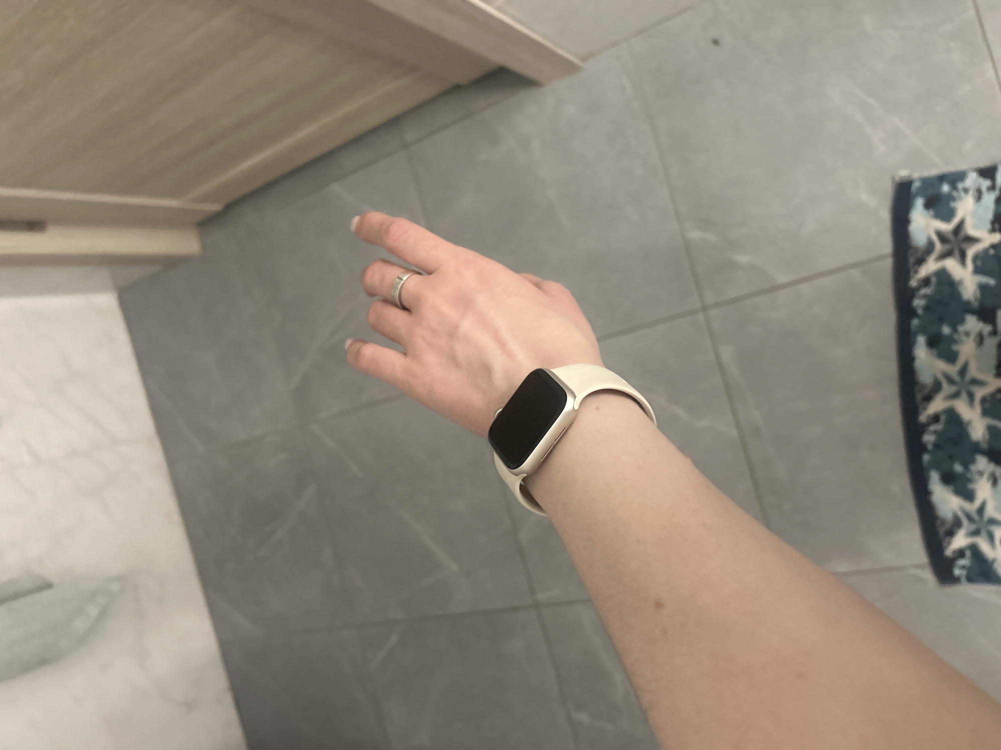The image shows a close-up view of a person's left arm and hand, wearing a white smartwatch and a silver ring. The arm is positioned horizontally across the frame, with the smartwatch on the wrist and the hand extended forward. The background consists of a tiled floor with a subtle marble pattern, and a wooden doorframe to the right. The scene appears to be indoors, possibly in a bathroom or hallway. There is no information available to determine the location in the city of Нижние Муллы, Russia.
