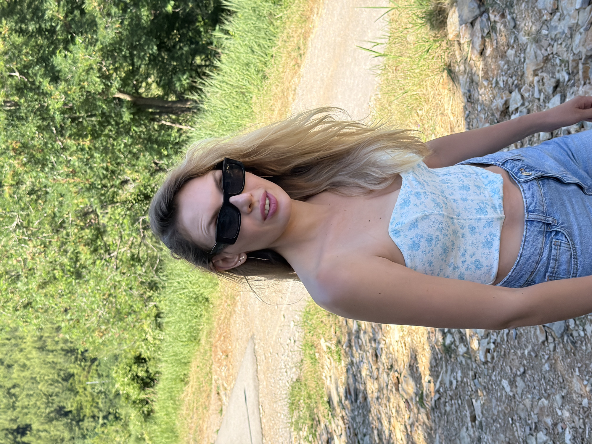 A young woman with long, light-blonde hair is captured in a medium shot, rotated 90 degrees clockwise, indicating she's likely standing outdoors. She is wearing dark sunglasses, a light blue and white floral strapless top, and light blue denim jeans. Her lips are slightly parted, revealing some of her teeth.
The background shows a natural outdoor setting with a dirt or gravel path that winds through green grass and scattered rocks. Lush green trees and foliage are visible further in the background. The lighting suggests it is daytime, and the shadows indicate a sunny day. The overall impression is casual and relaxed.
There is no visible text in the image.